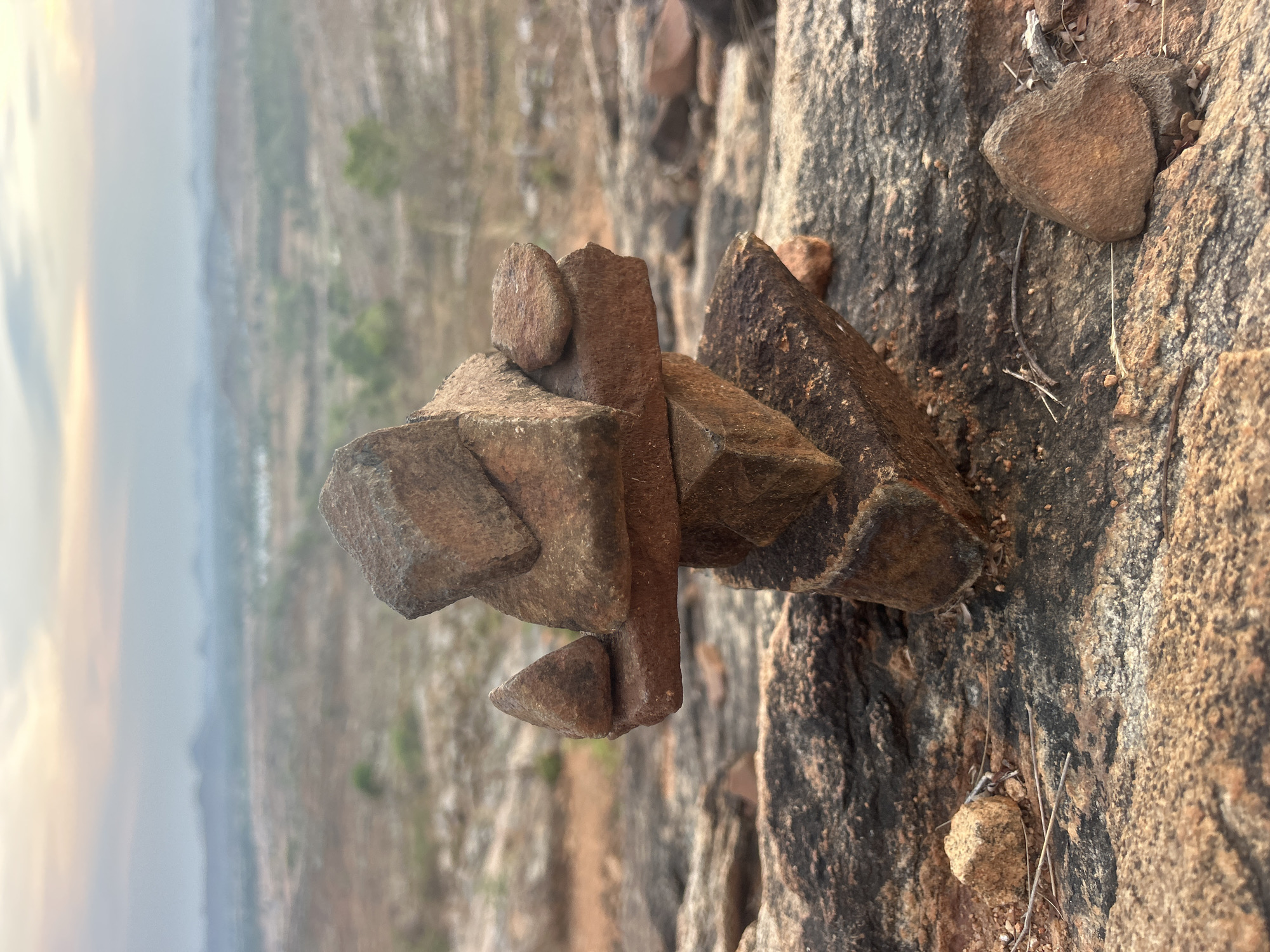 The image depicts a stack of rocks arranged artistically on a rocky surface. In the background, a hazy landscape is visible. The location is Kottala, India.