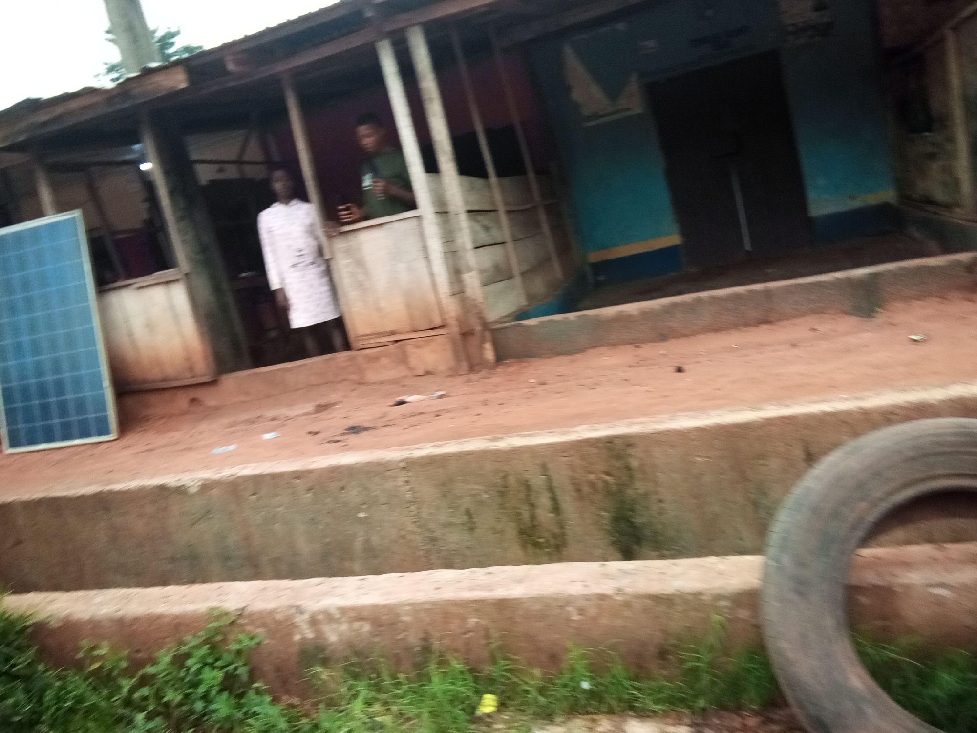 In Amaokpala, Nigeria, a solar panel is positioned to the left of a low-rise structure with an open front and wooden support beams. A person wearing a white patterned garment stands in the open doorway. To the right, a second person wearing a green shirt is visible inside the structure, holding a dark object, possibly a phone. The ground in front of the structure is reddish-brown earth, with a tire visible in the lower right corner of the frame. The structure itself appears aged, with wooden panels and a corrugated metal roof. The background is obscured by shadow and foliage.