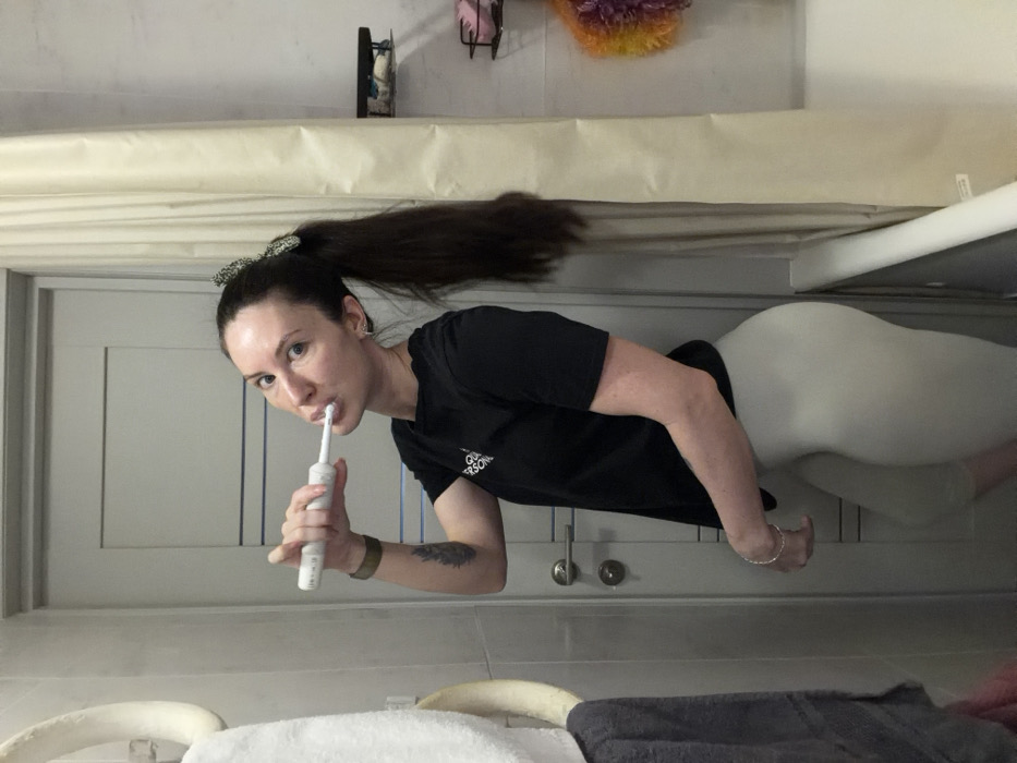 A woman with long brown hair pulled back in a high ponytail is brushing her teeth with an electric toothbrush in a bathroom. She is wearing a black t-shirt and light-colored leggings. The bathroom has gray cabinets and a white countertop. On the countertop are towels and a towel warmer. Above the cabinets are decorative items. The image is rotated, with the ceiling appearing on the left side.