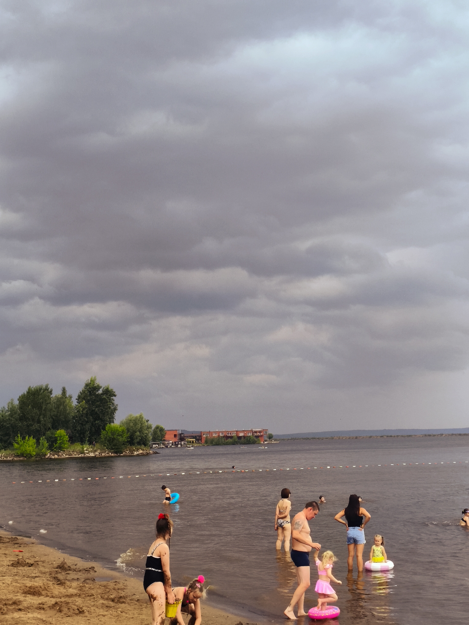 A group of people are enjoying a day at the beach, with some wading in the water and others playing on the sand. The scene is set against a dramatic sky filled with dark, ominous clouds, suggesting an impending storm or the aftermath of one. The water is calm, and a line of buoys marks a boundary in the distance.
In the foreground, two young girls are playing on the sandy shore. One, wearing a black swimsuit, is holding a yellow bucket, while the other, in a pink swimsuit, is standing on a pink inflatable ring. Further out in the water, a man in dark swim trunks is holding hands with a young girl in a pink skirt and swim top, who is also on a pink inflatable. Behind them, a woman in a black tank top and denim shorts stands with her hands on her hips, facing away from the camera. Another woman, in a patterned bikini, is further back in the water. A young boy in a black swimsuit is visible near a blue inflatable ring.
In the background, a treeline with lush green foliage stretches along the shore. Beyond the trees, a building with red roofs is visible, suggesting a developed area or resort. The overall atmosphere is one of a typical summer day at the beach, albeit with the subdued lighting and dramatic clouds creating a somewhat moody backdrop. The time of day is difficult to ascertain due to the cloud cover, but the general lighting suggests it is likely afternoon or late afternoon. There are no discernible emotions expressed by the people, as most are partially obscured or facing away. No visible text is present in the image.