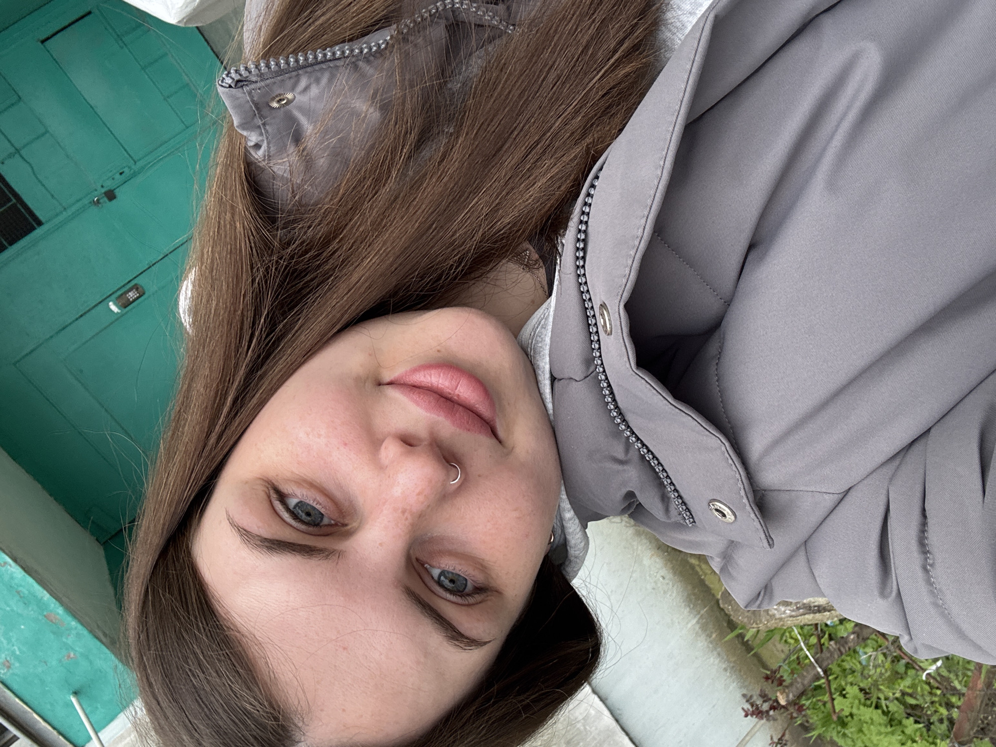 A young woman with brown hair and light blue eyes is taking a selfie. She has a nose ring on her left nostril. She is wearing a gray hooded sweatshirt and a gray puffy jacket. To the left of her is a teal door. To the right, there is concrete, grass, and small plants. There is no indication or basis to conclude any information about this image from Chornomorskoe, Ukraine.