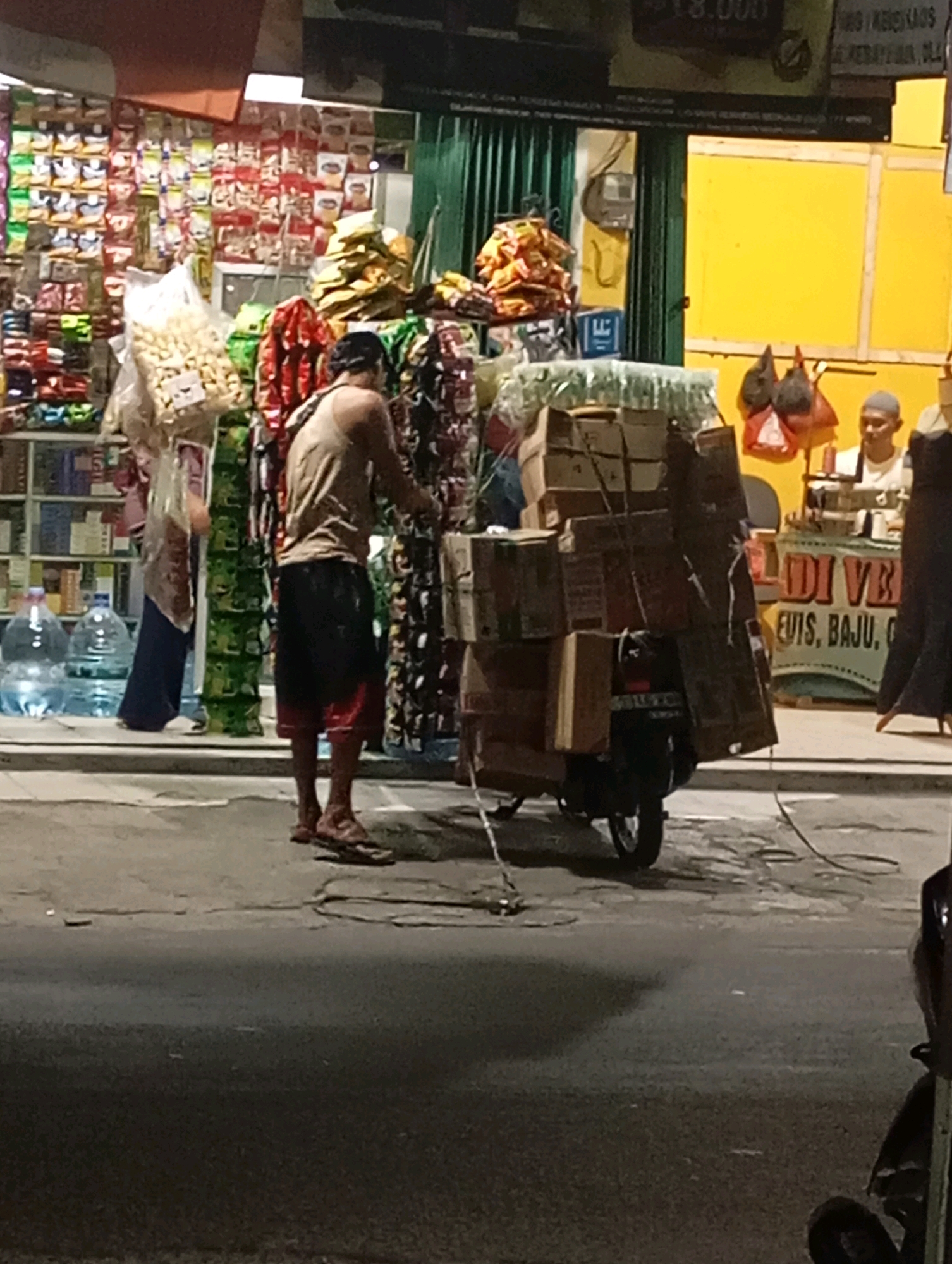In Babelan, Indonesia, a man wearing a tank top and shorts is loading goods onto a scooter. He is standing in front of a store, which appears to be a convenience store selling various products. The scooter is heavily laden with boxes and packages, suggesting a delivery or transport operation. The store's facade displays numerous shelves stocked with packaged goods, and large water bottles are visible outside. Another person, partially obscured, can be seen in the background, possibly working at the store. The overall scene depicts a common urban commercial activity in Indonesia, with a focus on street-level businesses and transportation.