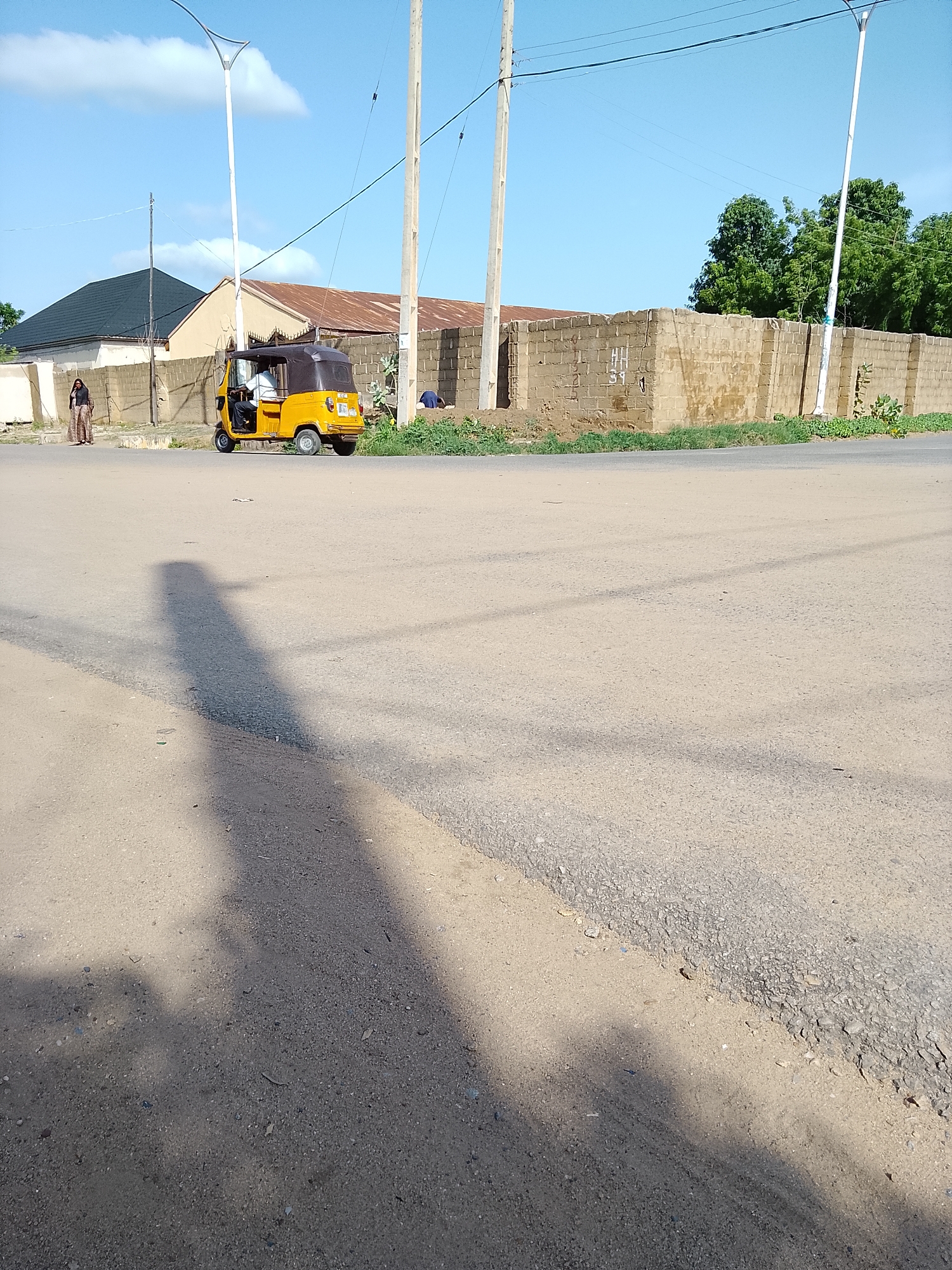 In Wauro Leggal, Nigeria, a yellow tricycle, commonly known as an "okada" or "keke," is stopped at a dusty intersection. The tricycle, with its black canvas roof, carries a driver wearing a white shirt and a passenger. A woman dressed in a patterned skirt and top stands on the side of the road near a brick wall. Behind the tricycle and the woman, buildings with corrugated metal roofs are visible. The environment is characterized by a paved road with a dirt shoulder, dry grass, and scattered trees in the background under a clear blue sky. A tall, modern street lamp stands near the road. The perspective is from ground level, capturing the tricycle and figures in the mid-ground, with a large shadow of the photographer stretching across the foreground.
