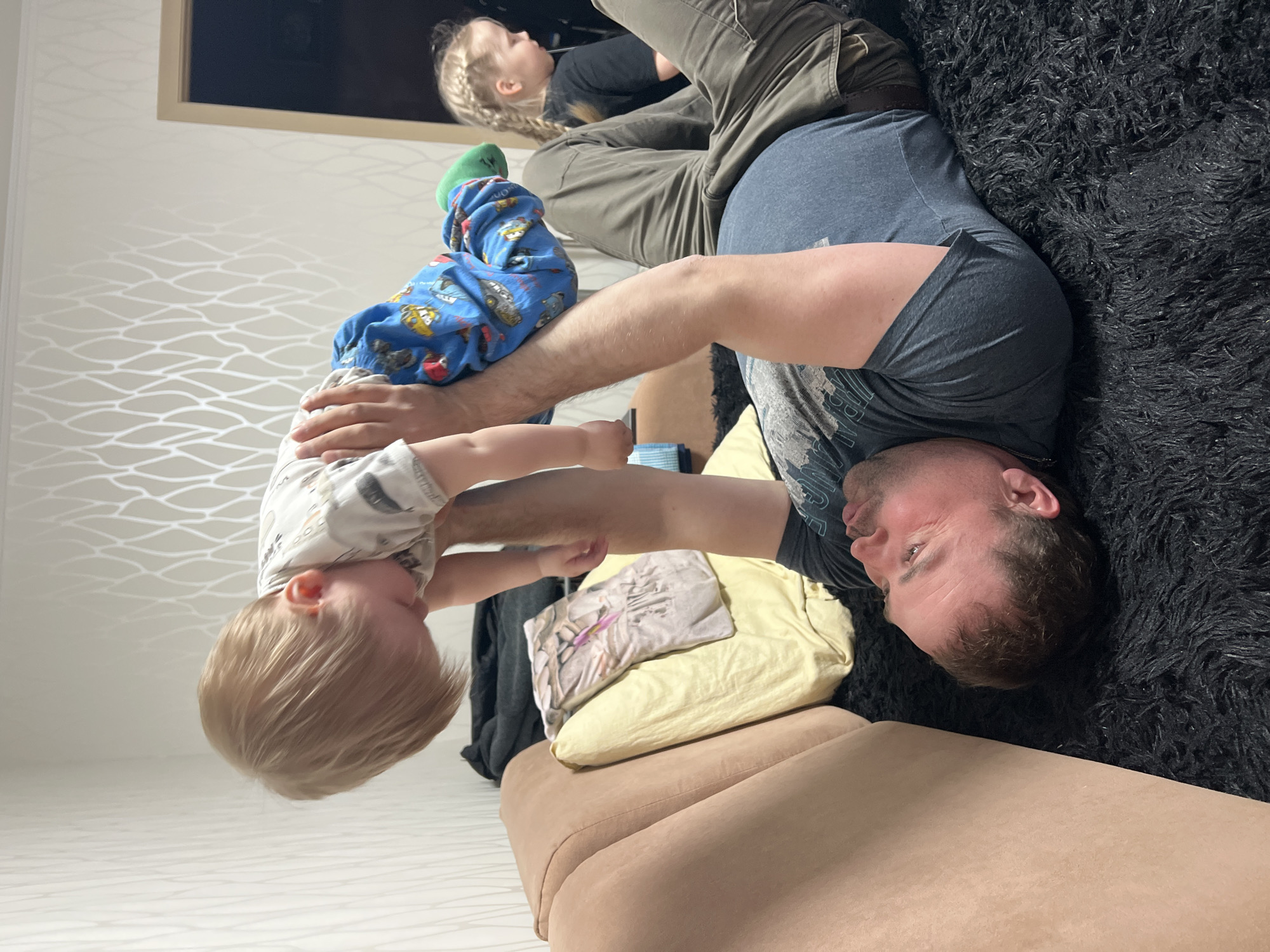 An indoor scene in Perm, Russia shows a man playing with two children. The man is lying on his back on a dark, shaggy rug, positioned next to a light brown upholstered couch. He has short hair and a beard, and is wearing a dark t-shirt. Above him, a toddler with light blonde hair is held upside down, likely by the man, in a playful interaction. The toddler is wearing a light-colored long-sleeved top, blue patterned pants with vehicle motifs, and green socks. Their head is hanging downwards, suggesting they are being swung or lifted in an inverted position.
In the background, another child with light braided hair is visible, lying on the light brown couch and looking upwards. This child is wearing olive green cargo pants and a dark top. On the couch next to the man are two pillows: one yellow and one with a subtle pattern. The wall behind them features light-colored wallpaper with a repetitive, wavy, geometric pattern. Part of a dark-framed object, possibly a picture frame or television, is seen mounted on the wall in the upper background. The lighting suggests an indoor setting, possibly during the daytime or evening, with no specific outdoor light cues. The overall mood appears to be one of playful family interaction. No discernible text is visible in the image.