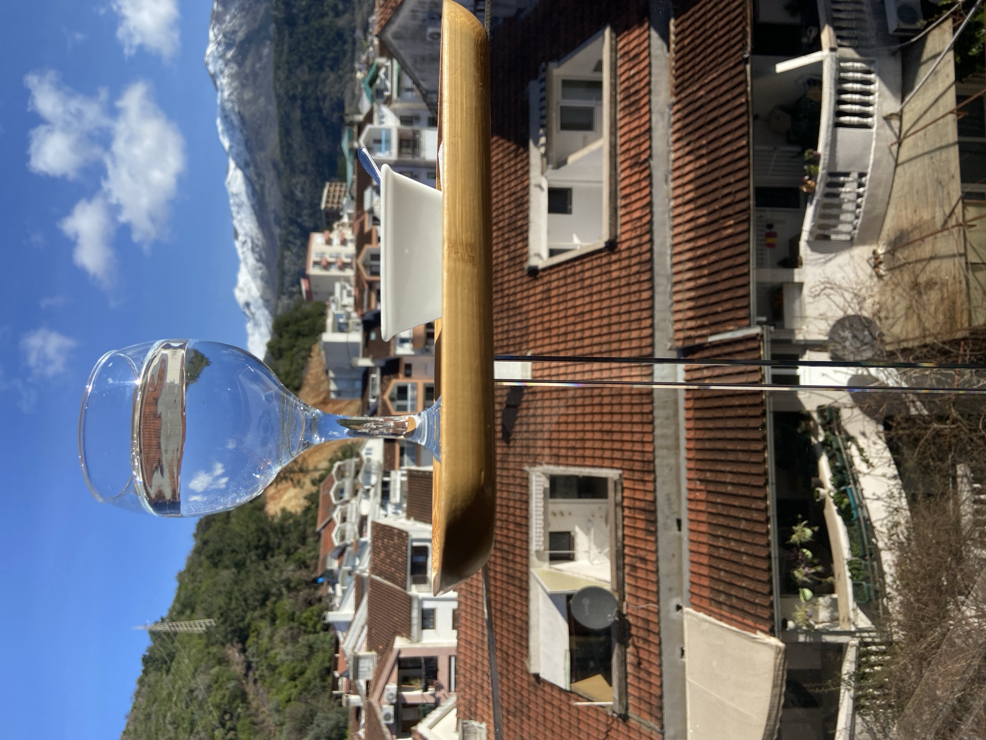 An outdoor scene in Budva, Montenegro, captured on a clear, sunny day. In the immediate foreground, a clear glass of water, approximately two-thirds full, stands on the left side of a horizontal, light-colored, oblong tray. To the right on the same tray, a small white ceramic bowl containing a spoon is visible. These items are supported by a slender, vertical metallic rod, suggesting placement on an elevated platform or balcony.
Below and extending into the midground is a dense residential cityscape composed primarily of multi-story buildings with white or light-colored facades and numerous red terracotta tiled roofs. Details observed include a dark satellite dish on one roof and balconies, some featuring white awnings and potted plants.
The background on the left reveals a steep, green, tree-covered hillside, surmounted by a prominent lattice-structure transmission tower. To the right, a panoramic view of majestic snow-capped mountains stretches across the horizon, contrasting with the bright blue sky and scattered white cumulus clouds.