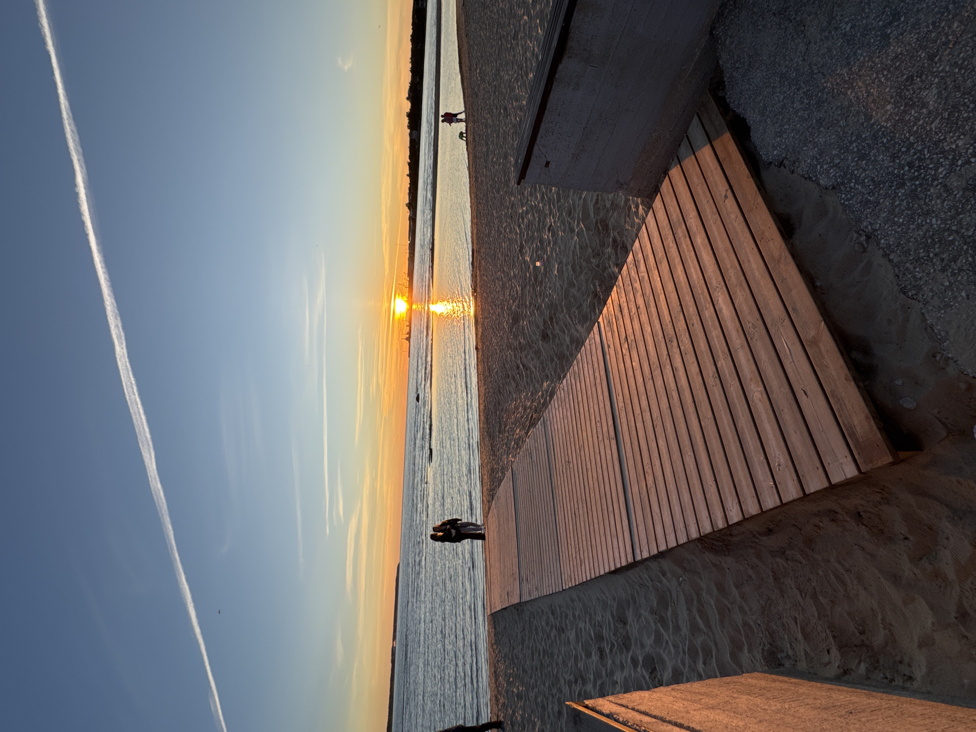 The image depicts a sunset over the Baltic Sea on a beach in Tallinn, Estonia. A wooden boardwalk extends from the foreground into the sandy beach, leading towards the water. Two people stand on the beach near the water's edge, facing the sea. In the distance, the sun is setting, casting a warm glow on the water and sky. A contrail from an airplane stretches across the blue sky, and a few scattered clouds are visible. The scene is serene and captures a moment of natural beauty at dusk. The specific location within Tallinn is likely a public beach area with amenities like boardwalks and seating.