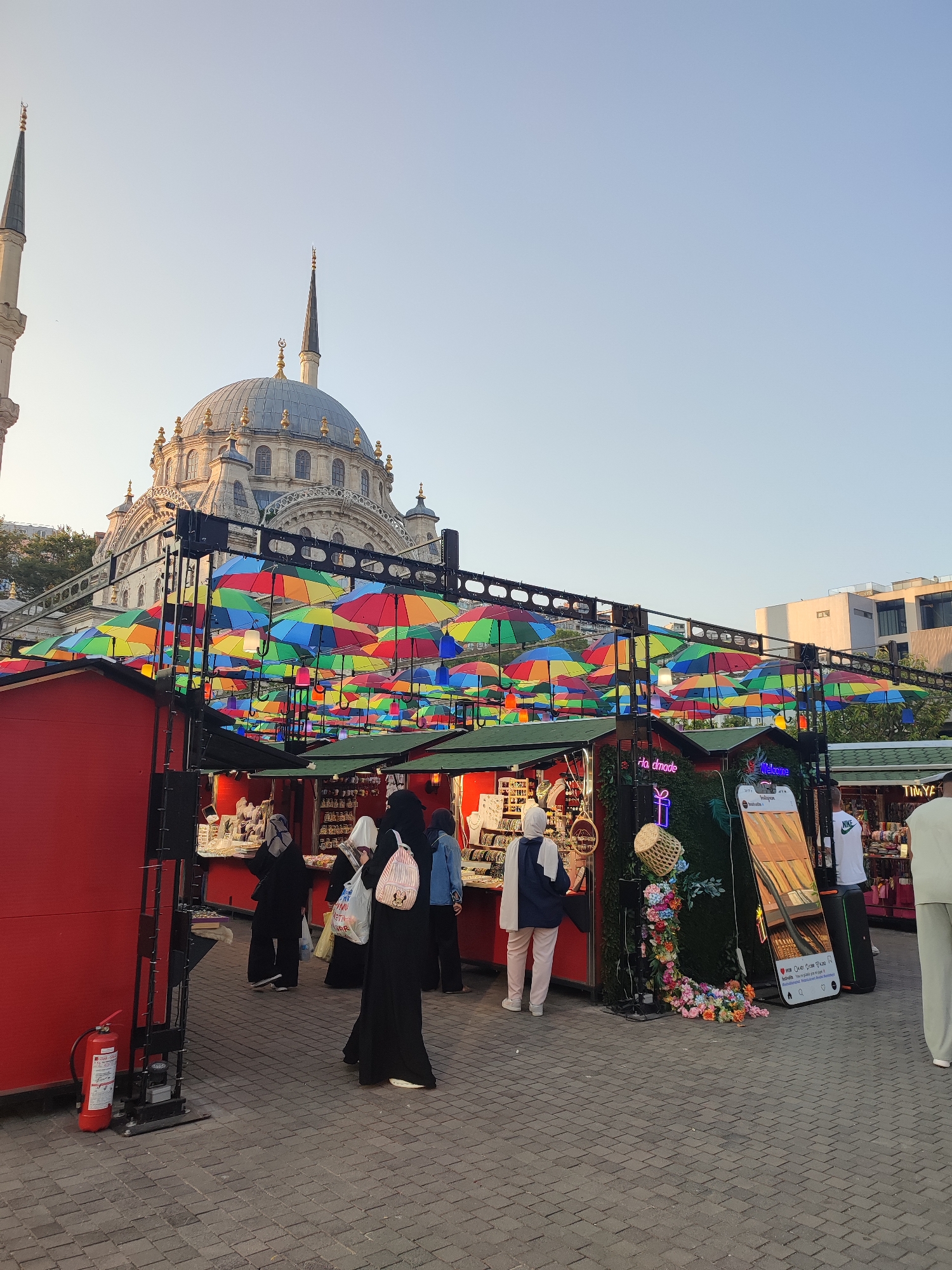 In Beyoğlu, Turkey, a market area is set up with stalls displaying various goods. Overhead, numerous colorful umbrellas are suspended, creating a vibrant canopy. A large mosque with a dome and minarets serves as a prominent backdrop to the scene. Several individuals, predominantly women wearing hijabs and abayas, are seen browsing the market stalls. One woman in the foreground is carrying a backpack with a Mickey Mouse design. A red fire extinguisher is visible near one of the stalls. The ground is paved with cobblestones. The overall atmosphere appears to be that of a lively outdoor market in a historic urban setting.