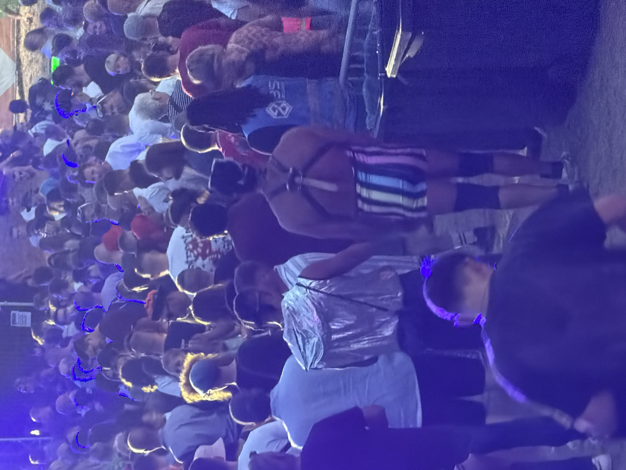 The image depicts a crowded outdoor event, likely a festival or concert, with a large gathering of people. The lighting is predominantly purple and blue, suggesting artificial stage lighting. In the foreground, a person wearing a silver vest and striped shorts is visible from the back, facing away from the camera. To their left, another individual in a light blue shirt is also seen from behind. Behind them, a large crowd of people are standing, many of them wearing casual clothing. The environment appears to be a gritty outdoor space, possibly with a dirt or gravel ground. There are some large dark structures or barriers in the background, one of which resembles a trash receptacle. The overall impression is of a lively, densely populated event. The specific location in Echternach, Luxembourg is not identifiable from the visual cues within the image.