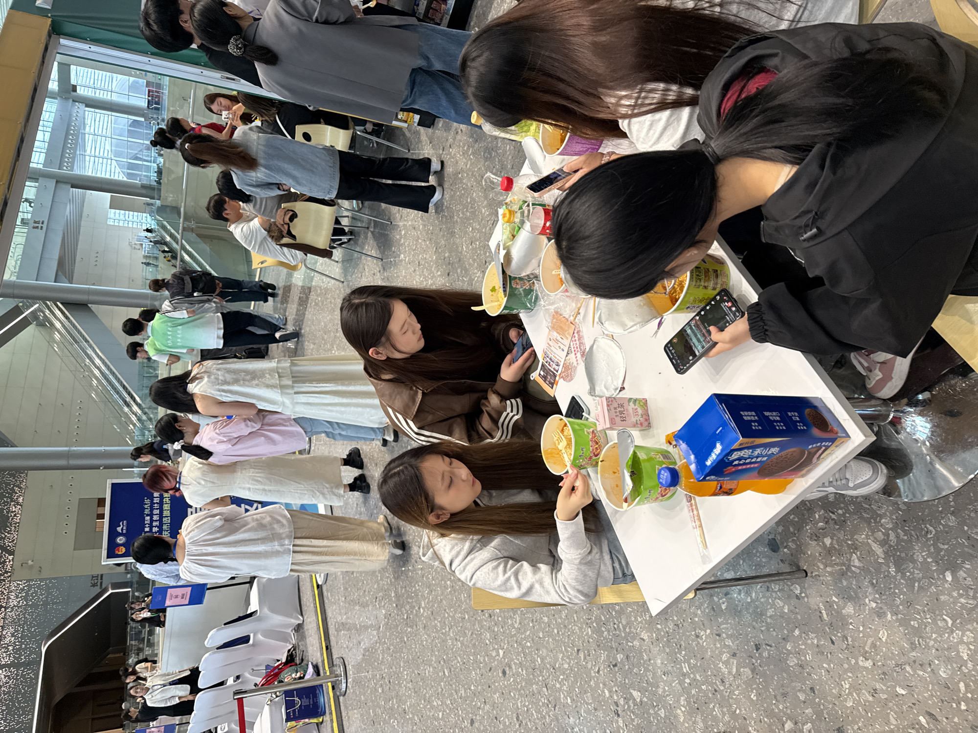 The image depicts a group of young people seated at a white table, engaged in eating instant noodles and interacting with their mobile phones. The setting appears to be an indoor public space with a polished grey speckled floor. In the background, escalators and architectural elements suggest a modern building, possibly a convention center or shopping mall. Several individuals are standing in the mid-ground, some appearing to be event staff or participants. One person in the foreground is wearing a black hooded jacket and holding a smartphone. Another person to their left, wearing a grey hoodie, is actively eating noodles. Other individuals at the table are also focused on their food or phones. A blue box of cookies and several bottles of beverages are visible on the table. The overall atmosphere suggests a casual gathering or a break during an event. The context provided indicates this scene is in the Chongqing High-tech Zone, China.
