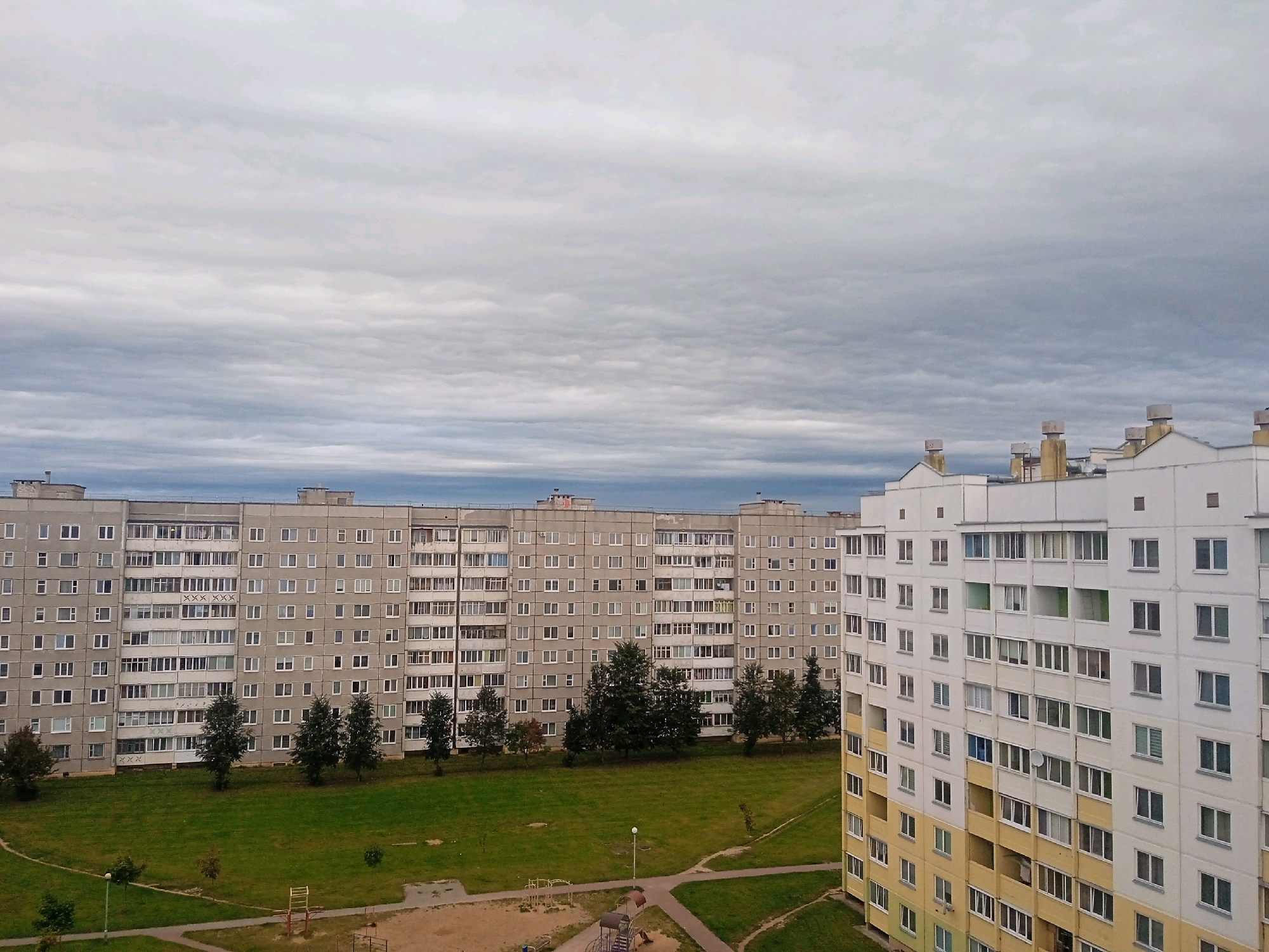 The image depicts a residential area in the city of Lida, Belarus, under an overcast sky. In the foreground, to the right, is a multi-story apartment building with a white facade and yellow accents on lower floors. It features numerous windows and balconies. To the left, and further back, is another large, grey apartment building with a repetitive design of windows and balconies. Between the buildings, a grassy courtyard is visible with several trees. In the lower left portion of the courtyard, playground equipment, including swings and a slide, is present. Paved pathways wind through the courtyard. The overall scene conveys a typical urban residential landscape.