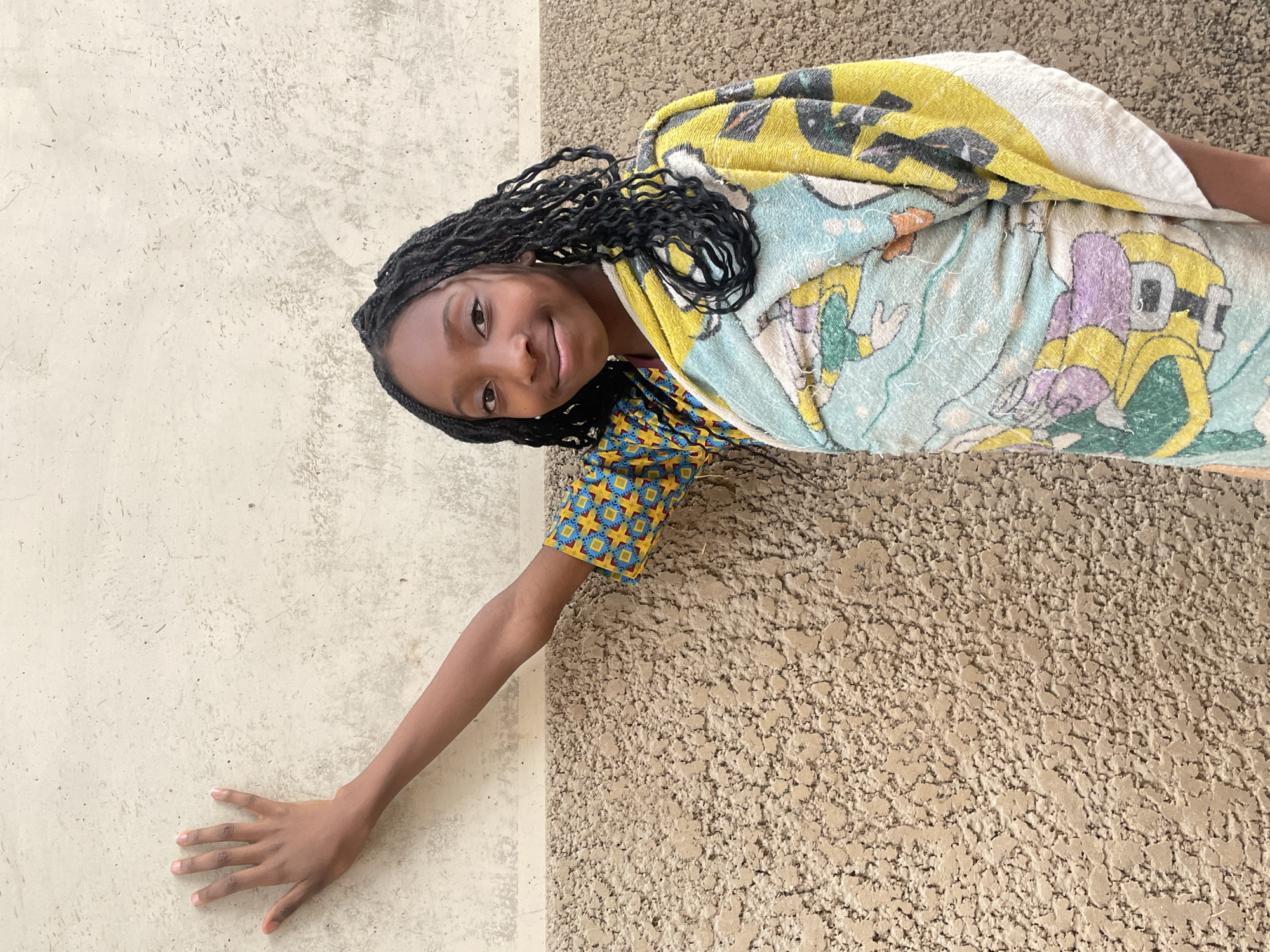 A young girl with dark hair styled in braids is pictured in a somewhat tilted perspective. She is partially wrapped in a towel that features cartoon characters, predominantly in yellow and blue. She is wearing a patterned top with blue, yellow, and red geometric shapes. Her right arm is extended towards a light-colored wall with a smooth texture. She is smiling and looking at the camera. The background is divided into two sections: the aforementioned light-colored wall and a rough, textured wall in a beige or tan color. The location is in the city of Jos, Nigeria.