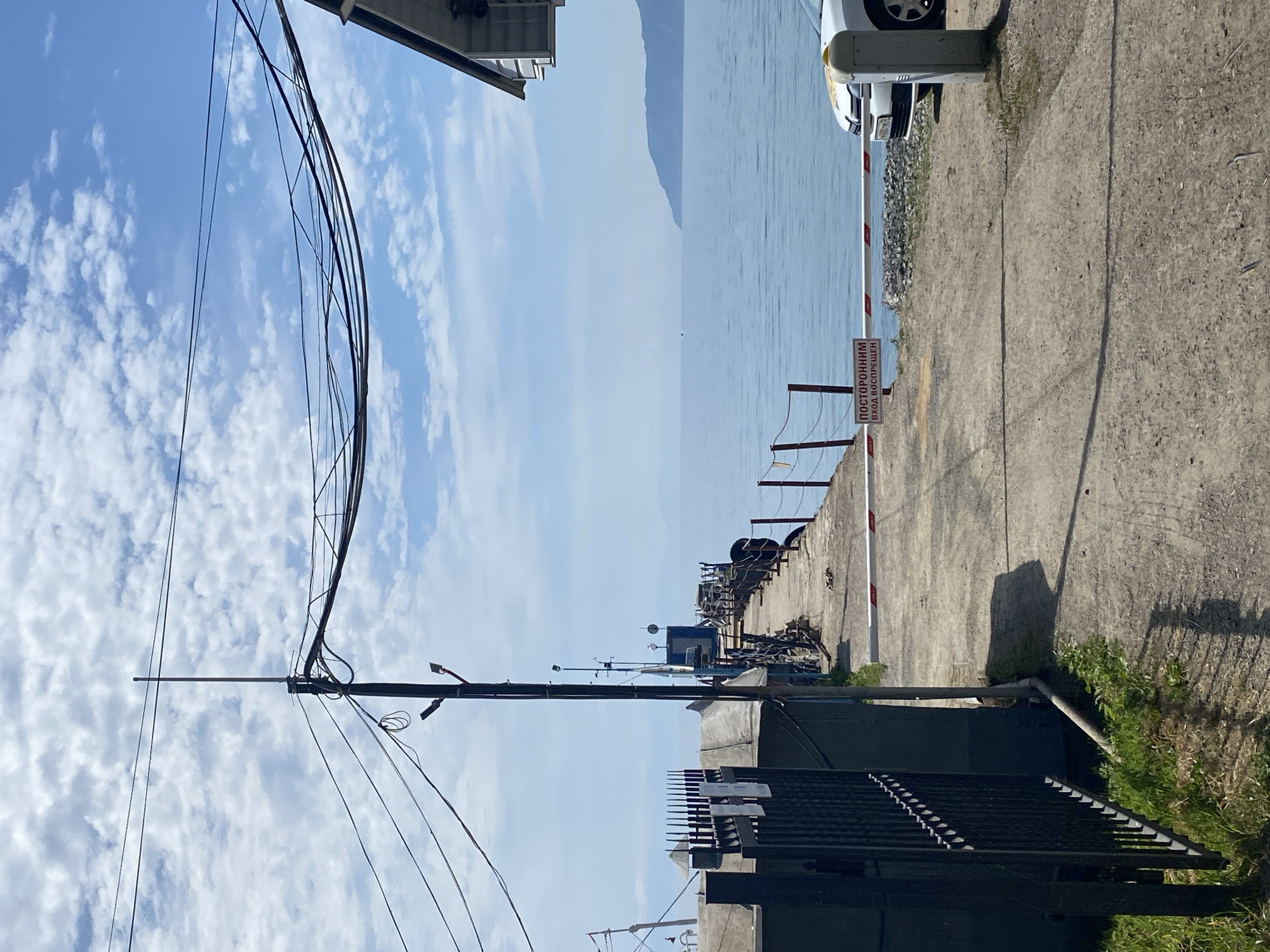 This is a photograph taken in Listvyanka, Russia, on a clear day. The scene depicts a concrete pier extending into a calm, blue body of water, likely Lake Baikal. Overhead, a complex network of electrical wires is strung between poles against a sky filled with scattered white clouds.
On the left side of the image, a black metal fence encloses a dark-colored structure, possibly a building or shed, with a peaked roof. Green grass grows along the base of the fence and structure.
The pier itself is a wide, concrete path leading out to the water. Along the right edge of the pier, a railing is visible, with mooring posts and thick ropes. Several small lights are mounted on poles along the pier. At the end of the pier, a blue building or container is situated.
A red and white striped barrier arm extends across the pier, blocking entry. A sign with red lettering on a white background is attached to the barrier arm, reading "ПОСТОРОННИМ ВХОД ВОСПРЕЩЁН" (Unauthorized entry prohibited). To the right of the barrier, a white car is parked, partially visible. The ground leading up to the barrier is a gravel and dirt surface.
In the distance, the horizon is marked by a hazy, mountainous shoreline. The overall impression is one of a functional port or docking area on a tranquil summer day.