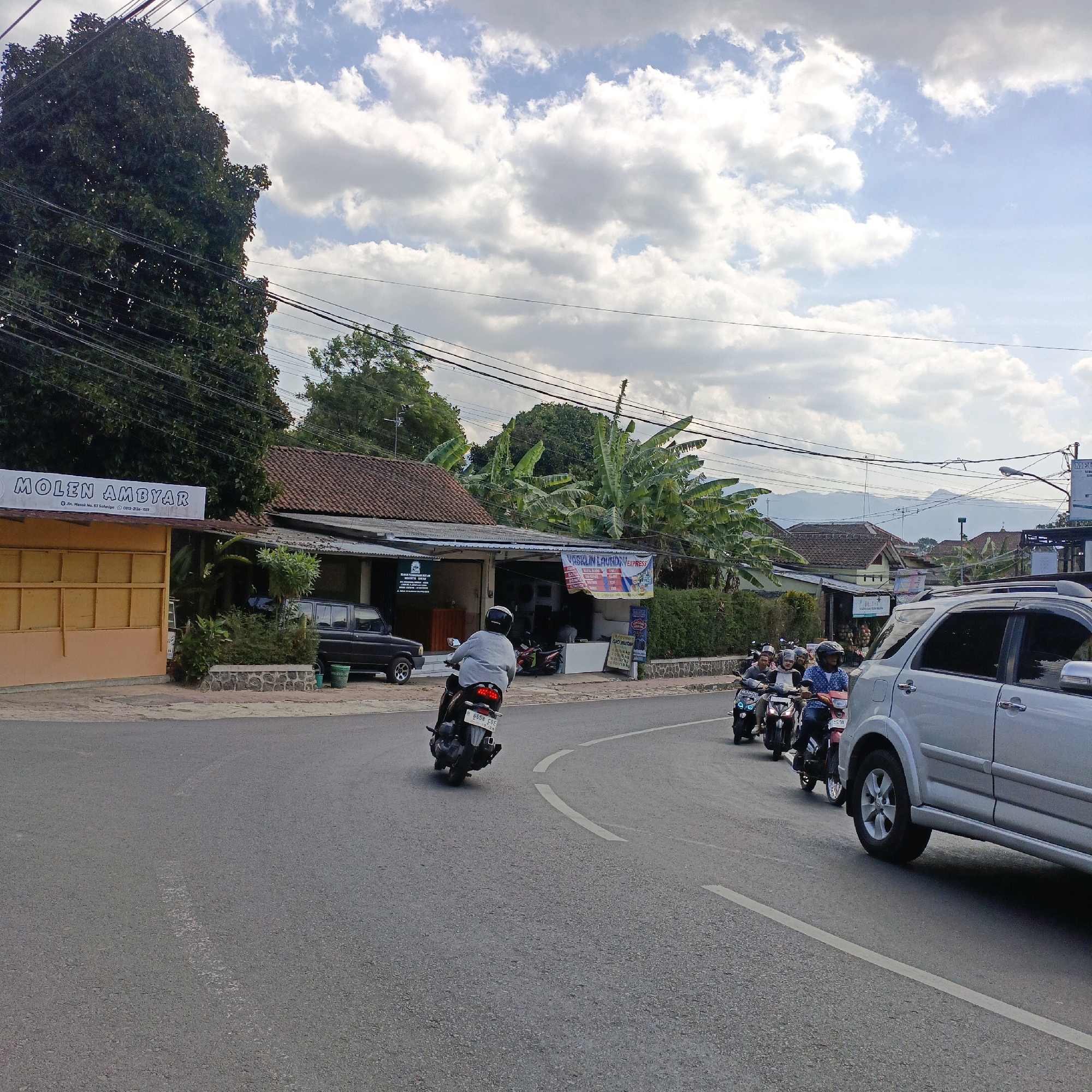 This image depicts a street scene in Salatiga, Indonesia, on a cloudy day. The road is paved with asphalt and features white lane markings. On the left side of the road, there is a building with a sign that reads "MOLEN AMBYAR" and below it, "Jln. Merok No. 53 Salatiga" with a phone number. Adjacent to this building are more structures with tiled roofs, some adorned with posters. A dark-colored van is parked near the buildings.
Several people are on motorcycles, either riding or parked. One motorcycle rider in the foreground is wearing a grey shirt and a white helmet, heading away from the camera. Further down the road, a line of motorcycles with riders is proceeding in the same direction. On the right side of the road, a silver SUV is driving away from the camera.
The environment includes lush green trees, including banana trees, and in the background, distant hills or mountains are visible under a sky filled with white, puffy clouds. Power lines are strung across the scene, supported by poles. The overall impression is a typical street in a bustling town in Indonesia, with everyday transportation and commerce taking place.