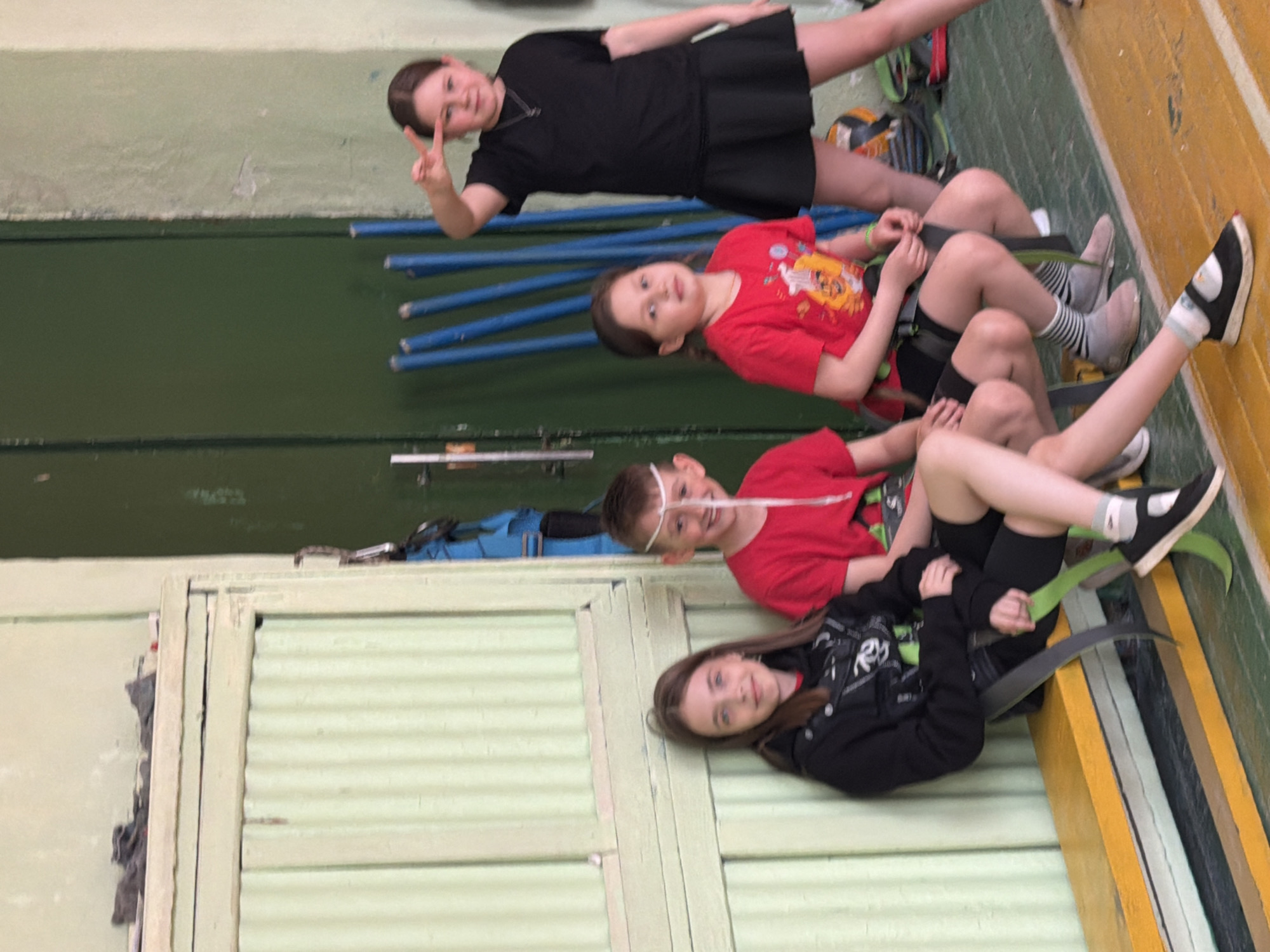 Four children are pictured indoors, likely in a sports hall or gymnasium in Izhevsk, Russia, seated on what appear to be custom-made boards or sleds. The image is oriented sideways, but if rotated upright, the children are arranged horizontally.
From left to right:
The first child, a girl, wears a black short-sleeved top and a black pleated skirt. She is smiling and holding up two fingers in a peace sign.
Next to her is another girl wearing a red t-shirt with a graphic design. She is also smiling.
The third child is a boy, wearing a red t-shirt. He has a white string or band stretched across his face, passing over his nose. He is smiling.
The fourth child, a girl, is on the far right, wearing a black jacket with white graphics and black leggings. She is smiling and looking towards the camera.
The children are seated on individual, dark-colored sled-like structures with green or dark grey elements, some of which rest on a light-colored floor with yellow markings. In the background, there are blue horizontal bars, suggesting gym equipment, and walls of varying colors including dark green and a lighter, corrugated green paneling. Behind the leftmost girl's feet, a backpack, a helmet, and a volleyball are visible on the floor, indicating a sports-related context. All the children appear happy and engaged. There is no visible text in the image.