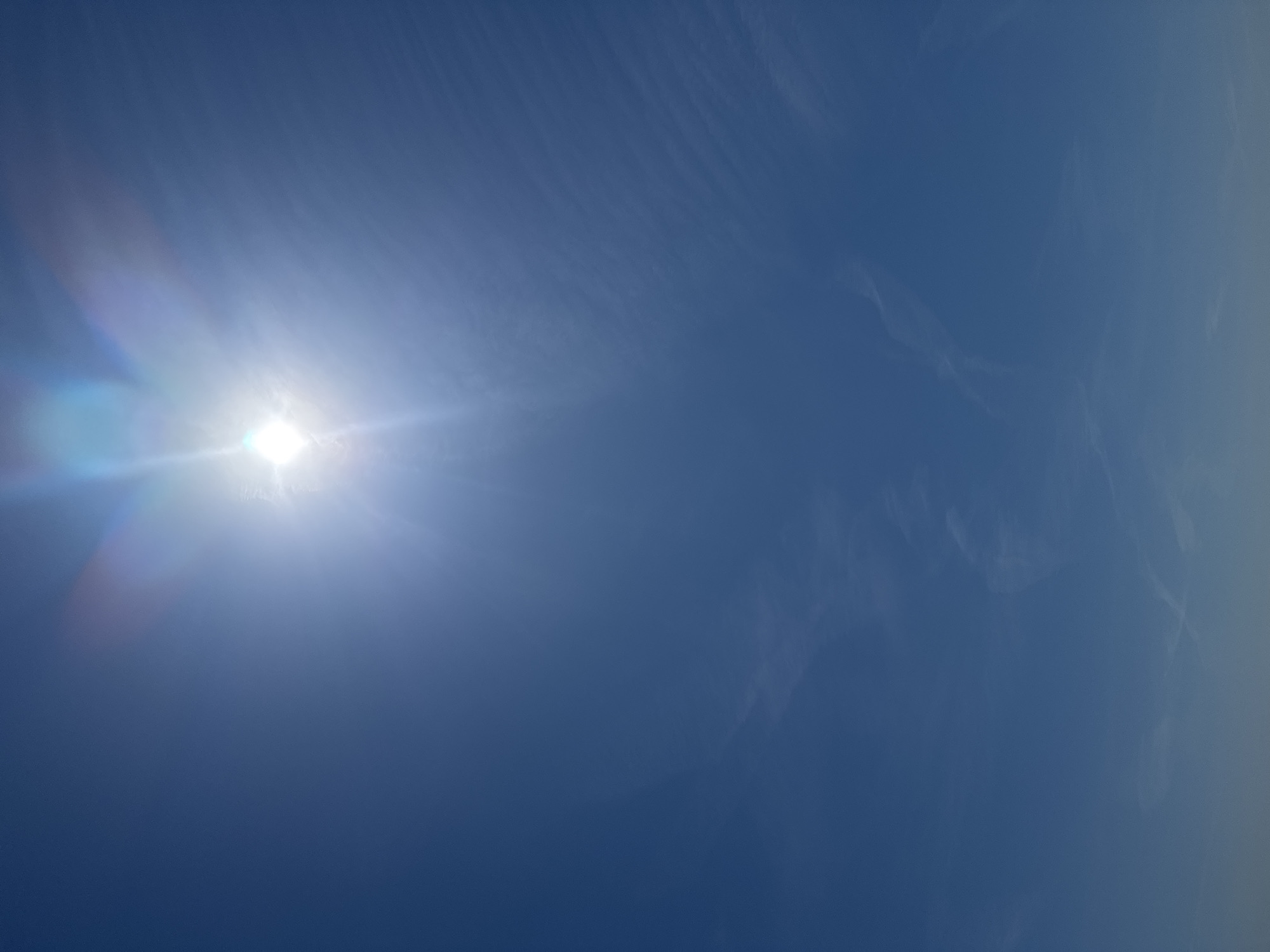 The image captures a vibrant blue sky illuminated by the sun, likely over Figueira da Foz, Portugal. The sun is prominently featured in the upper left quadrant, appearing as an intensely bright, overexposed white orb. Strong lens flares and distinct sun rays emanate from the sun, casting a luminous glow and exhibiting rainbow-like chromatic aberration across the left side of the frame. The sky is a rich, deep blue, subtly transitioning to a slightly lighter, perhaps hazy, tone towards the right edge of the image. Delicate, wispy white clouds, characteristic of cirrus or cirrostratus formations, are faintly visible, particularly in the upper and mid-right sections of the sky, forming ethereal, streaky patterns. These high-altitude clouds do not significantly impede the sun's brilliance. The scene suggests a bright, clear day, likely during the late morning or early afternoon. No people, man-made objects, or visible text are present in the photograph, which focuses entirely on the natural elements of the sky.