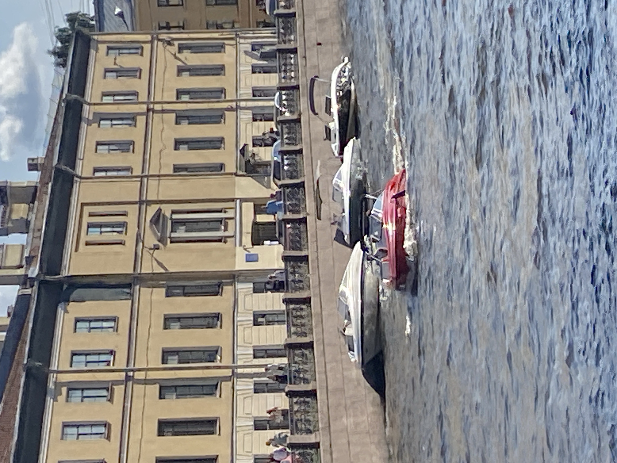 This is a medium-distance, eye-level, outdoor, sunny, daytime photograph of a canal in Saint Petersburg, Russia. In the foreground, three motorboats are visible on the water. The closest is a small, red, open-topped boat moving to the right. Behind and to the left of the red boat is a white motorboat with a canopy. To the right of the white boat and further back is another white motorboat with a dark hull and a dark canopy. The boats are moving on a body of water that appears to be a canal, given the presence of a stone embankment and a retaining wall.
Behind the canal, a row of yellow buildings with a dark green roofline stretches across the frame. The buildings have multiple windows, some of which are dark, and decorative architectural details. A black metal railing with an intricate design separates the embankment from the buildings. Several people are standing behind the railing, observing the canal. They are casually dressed, suggesting a tourist or leisure activity. The sky above is partly cloudy with blue patches. The overall scene depicts a tranquil urban waterway with recreational boats and historic architecture characteristic of Saint Petersburg.