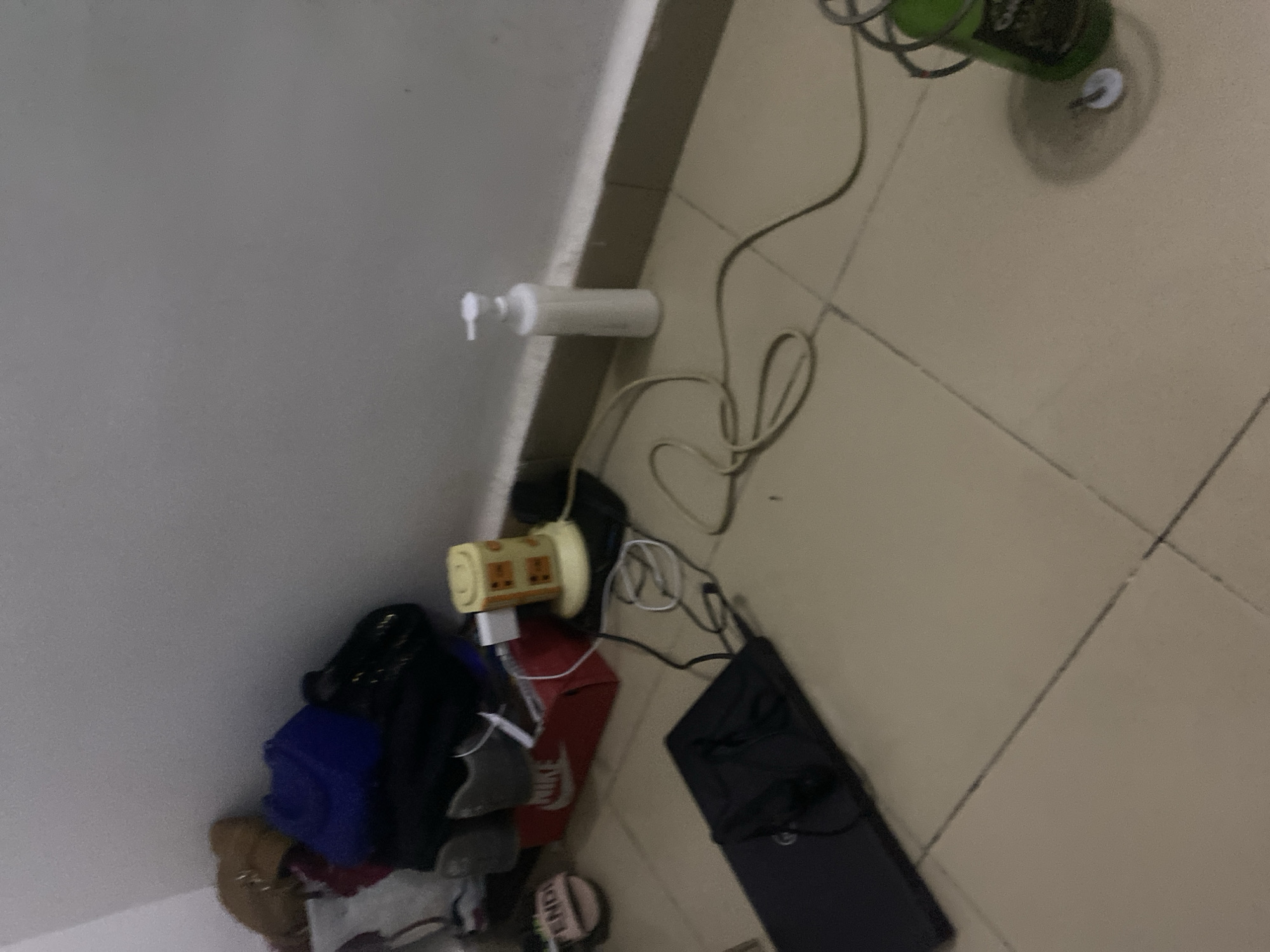 A cluttered corner of a room, likely residential, with items on the floor and stacked against the wall. The floor is tiled with a neutral beige color, and the wall is painted white.
In the center of the scene, a black laptop is open on the floor, with its screen facing away from the viewer and a black cable connected to it. To the left of the laptop, a pair of black earbuds is visible.
Behind the laptop, a red shoebox is placed against the wall, with the word "NIKE" printed on it in white letters. On top of the shoebox, two grey sneakers are stacked.
Further back and to the left, a pile of bags and clothing is visible. This includes a dark blue bag, a lighter colored bag, and what appears to be a brown handbag or purse on top.
To the right of the shoebox and sneakers, a small, yellow, cube-shaped electrical device with orange markings is plugged into a white adapter. Wires from this device are spread across the floor.
To the right of the yellow device, on the floor near the wall, is a tall white bottle with a pump dispenser.
In the far right of the image, a green bottle with a label that includes some text and a white cap is standing on the tiled floor. A small white object, possibly a cap or button, is located near the base of this bottle.
The scene appears to be indoors, possibly a bedroom or storage area, with a focus on the arrangement of personal belongings and electronic devices. There is no indication of the specific location within Howayi, Nigeria.