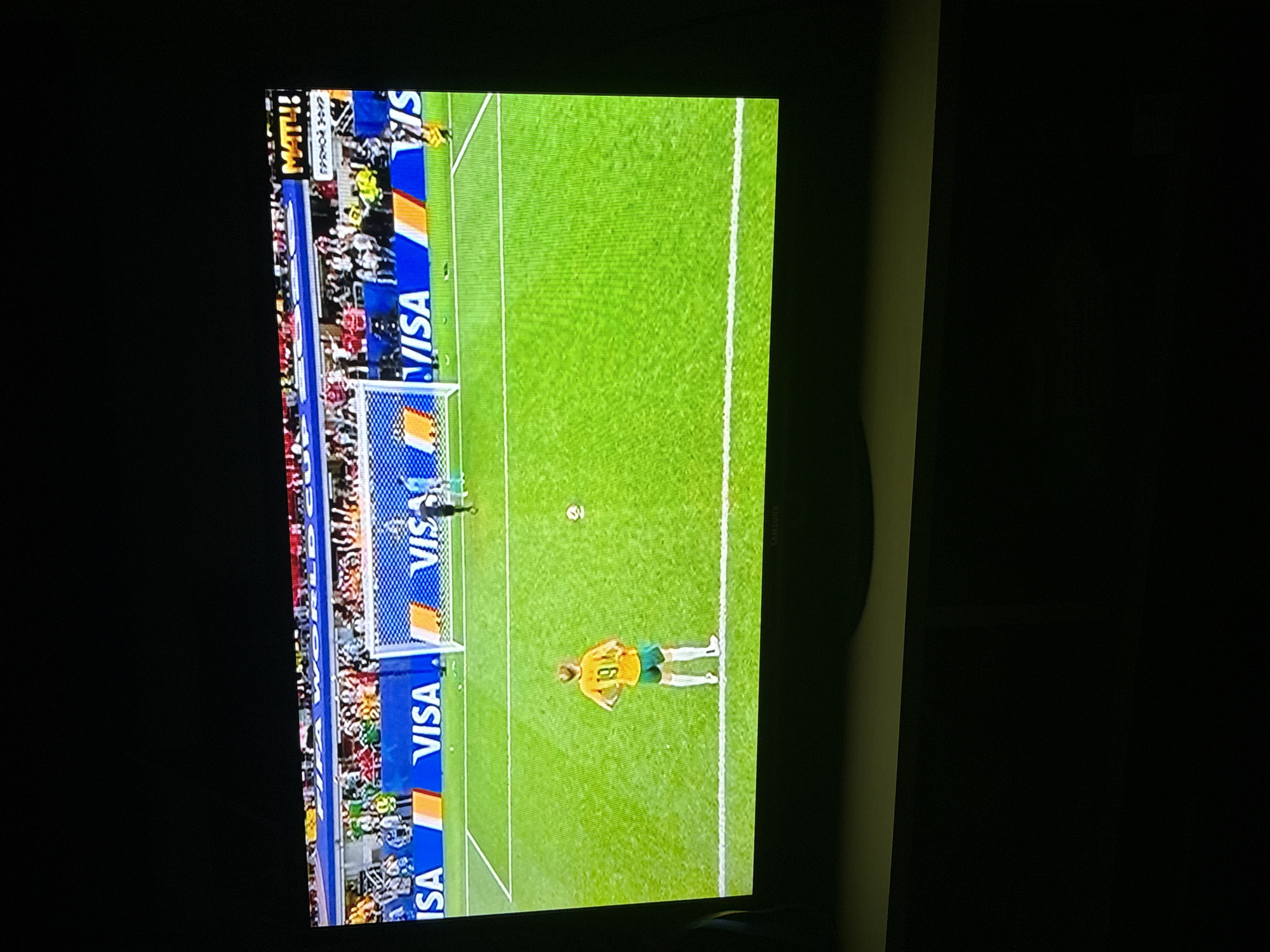 This is a television screen displaying a soccer match. The screen shows a soccer player in an orange jersey with the number 19 on the back, standing on a green field. There is a goal net and several other players in the background. The screen also displays advertising banners with the VISA logo. The top right corner of the screen shows "MATH!" in yellow letters. The location provided for this image is Butovo, Russia, however, there is no visual information within the image to confirm or deny this location.
