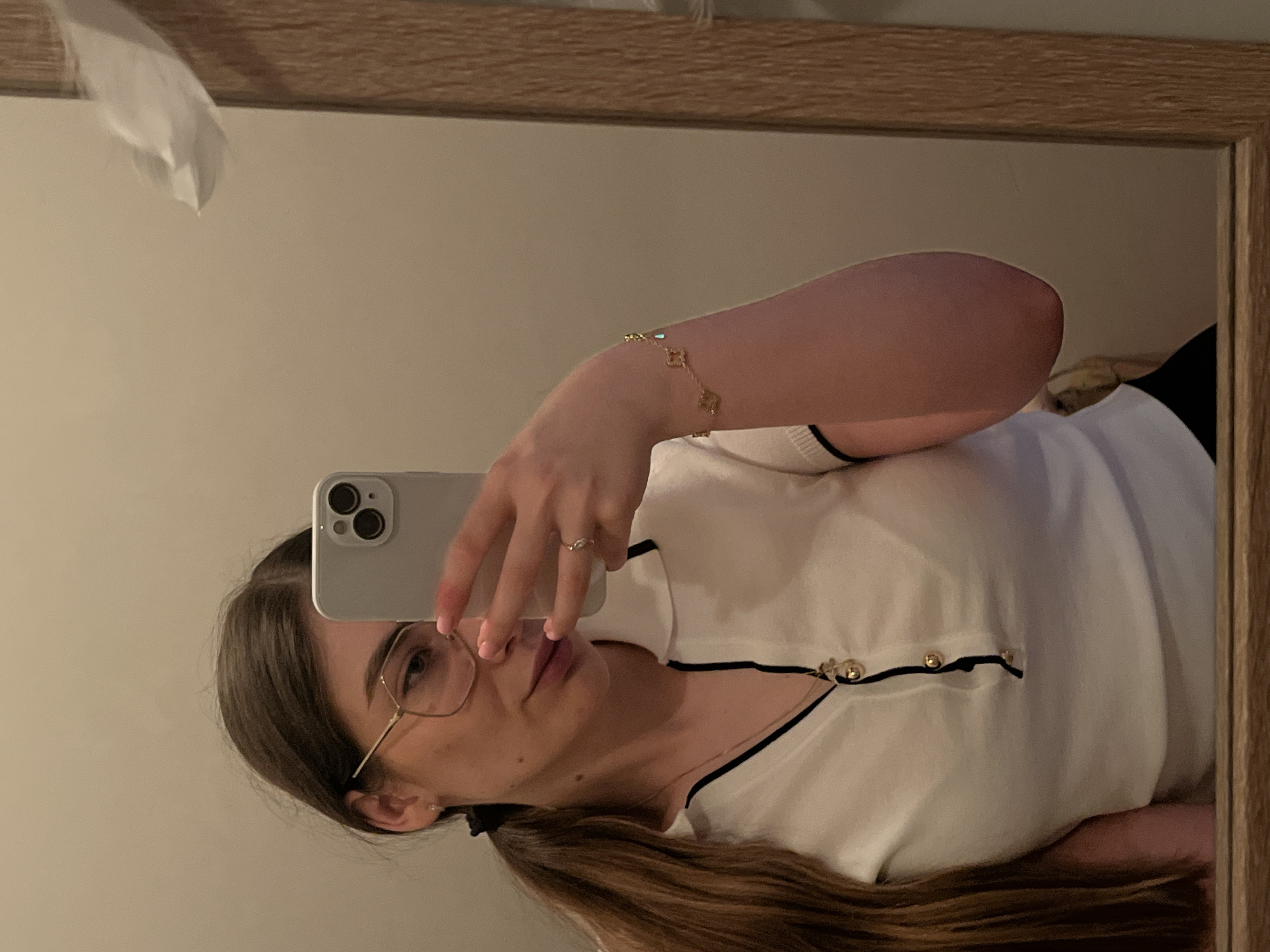 An indoor, waist-up mirror selfie captures a young woman in the city of Unknown, Unknown. The woman has long, straight dark brown hair pulled back into a low ponytail secured with a black elastic. She wears gold-framed hexagonal glasses. Her left hand, adorned with a delicate ring on the ring finger and light pink nail polish, holds a light gray smartphone, partially obscuring the right side of her face. She has a small stud earring in her visible left ear. She is wearing a white collared short-sleeved top with black trim along the collar and a black placket featuring three prominent gold-toned buttons. A long gold-toned necklace with a clustered charm pendant hangs around her neck. On her visible right wrist, she wears a thin gold-toned bracelet with small, regularly spaced decorative elements. The background, visible in the mirror, consists of a plain, light beige wall. A crumpled piece of white paper or tissue is visible in the upper right corner, partially obscured by the reflection. The mirror itself is framed by light-colored wood, visible on the right and bottom edges. The lighting is soft and even.