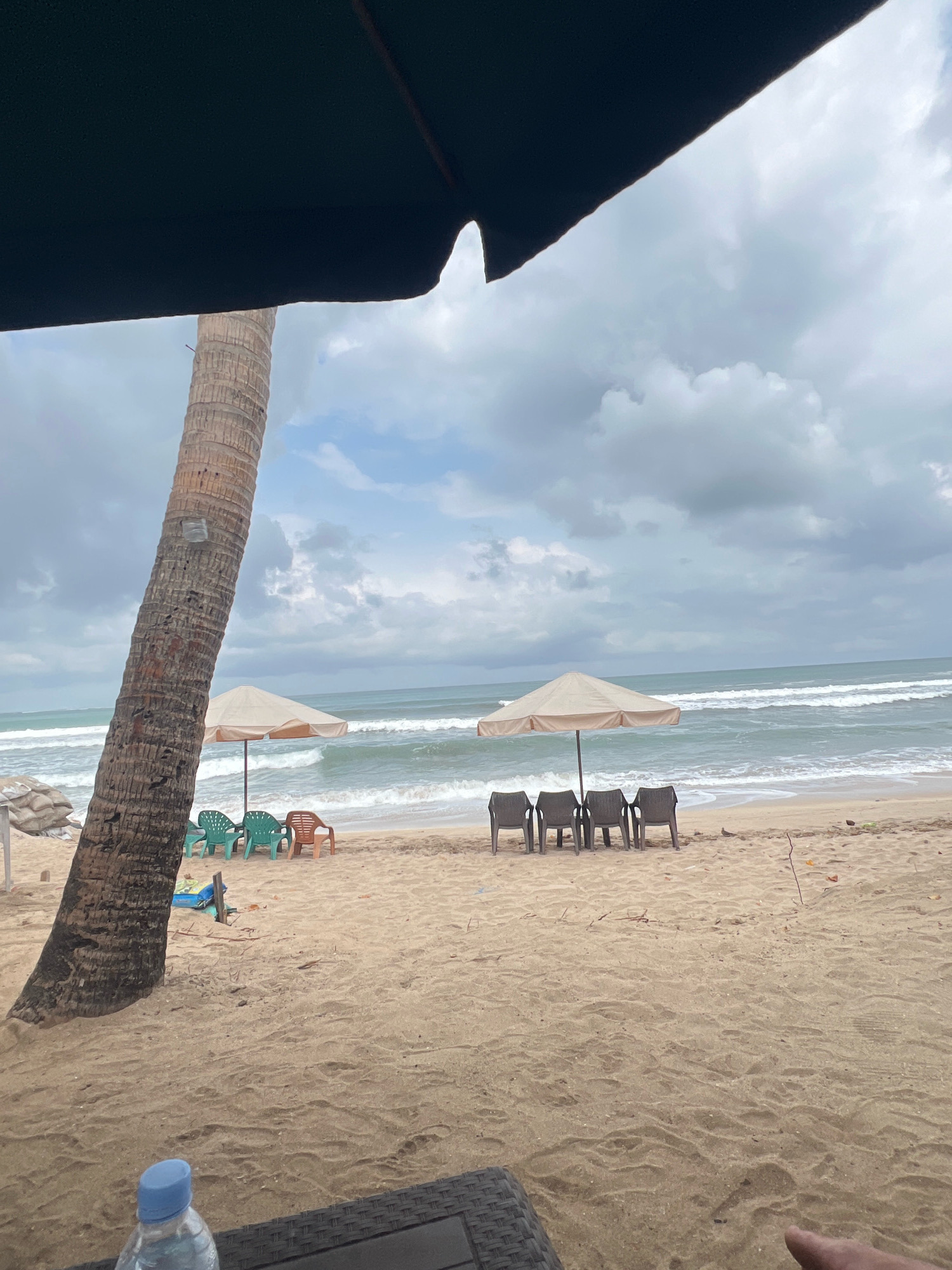 The image depicts a beach scene in Kuta, Indonesia, under a cloudy sky. A tall, textured palm tree trunk is prominent on the left side, partially obscured by a dark awning. In the midground, a sandy beach stretches towards the ocean, where gentle waves are rolling in. Two beach umbrellas are set up: one smaller one with three green chairs and one brown chair positioned in front of it, and a larger one with four dark chairs lined up before it. The sand is light brown and appears somewhat disturbed, with scattered small debris. In the foreground, a bottle of water with a blue cap rests on a dark, woven surface, suggesting a seating area. The overall atmosphere is one of a slightly overcast day at a tropical beach.