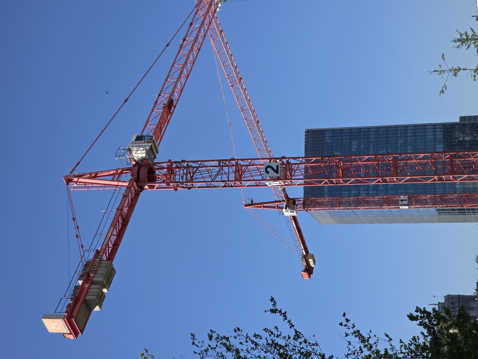 A red tower crane, possibly located in Warsaw, Poland due to the architectural style of the building behind it, is angled upwards against a clear blue sky. The crane has a counterweight on one end and a jib with a trolley on the other. A building with a glass facade is visible behind the crane. Tree branches with green leaves are in the lower portion of the frame. A small dark spot, possibly a bird, is visible in the sky. The number "2" is visible on the jib of the crane.