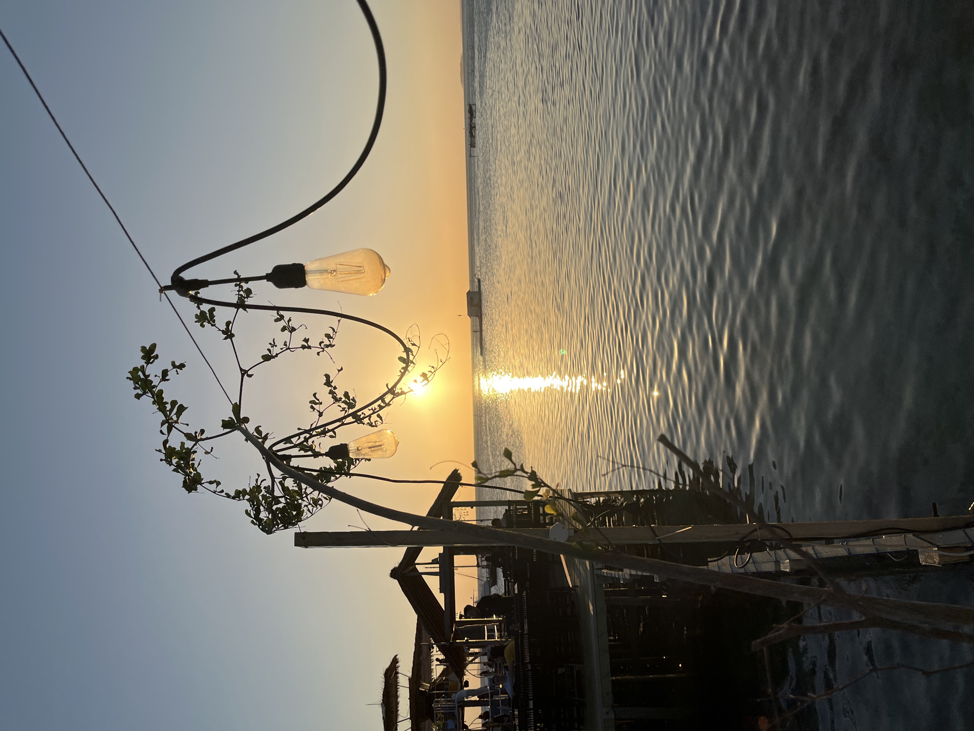 This is a photograph taken at sunset in Karimunjawa, Indonesia, capturing the view from a wooden pier. In the foreground, a string of incandescent bulbs hangs from a wire, casting a warm glow against the darkening sky. A vine with green leaves is intertwined with the wire and a wooden post, adding a natural element to the scene.
To the left, a section of a wooden pier is visible, with several thatched-roof structures that appear to be part of a restaurant or seating area. Silhouetted figures of people can be seen seated at tables within these structures, suggesting a relaxed dining or social gathering.
The main focus of the image is the calm sea and the setting sun. The sun, a bright orb, is positioned just above the horizon, its reflection creating a shimmering golden path across the water. The sky is a gradient of soft blues and oranges, typical of a sunset. In the distance, several small structures or boats are visible on the water, adding depth to the seascape. The overall atmosphere is tranquil and serene, characteristic of a tropical evening.