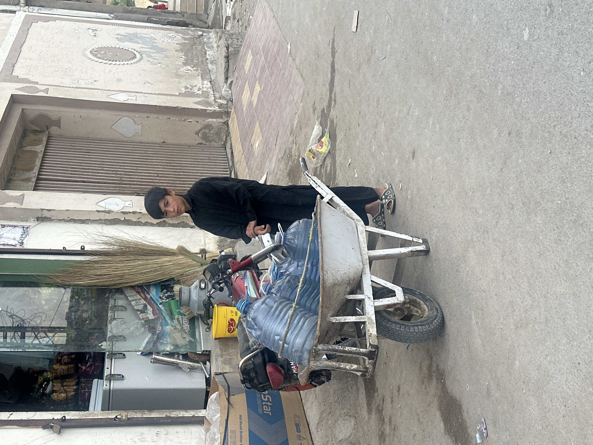 A young male in dark clothing pulls a metal cart filled with water bottles on a paved street in Quetta City Tehsil, Pakistan. The cart is attached to a red and grey motorcycle. A building with a closed metal gate and decorative designs is visible on the left side of the frame, next to a storefront with a glass window displaying merchandise. A cardboard box with "STAR" printed on it sits near the motorcycle. The street shows signs of wear and litter.