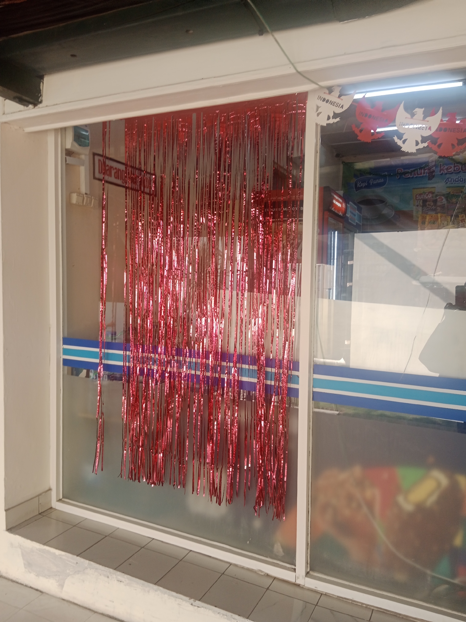 A red tinsel curtain hangs in the doorway of a convenience store. The store's exterior features a white frame around its glass windows and door. To the left of the curtain, a sign reads "DILARANG MEROKOK" (No Smoking). Decorations resembling the Indonesian national symbol, Garuda Pancasila, are visible in the upper right corner of the window, some in white and some in red, with the word "INDONESIA" written on them. Inside the store, shelves stocked with goods are visible, including advertisements for products such as "Kopi Panas" (Hot Coffee) and "SunCo". The store appears to be located in a commercial area, possibly on a street with a tiled sidewalk and a low concrete barrier in front of the entrance. The overall impression is of a store decorated for a national holiday or event.