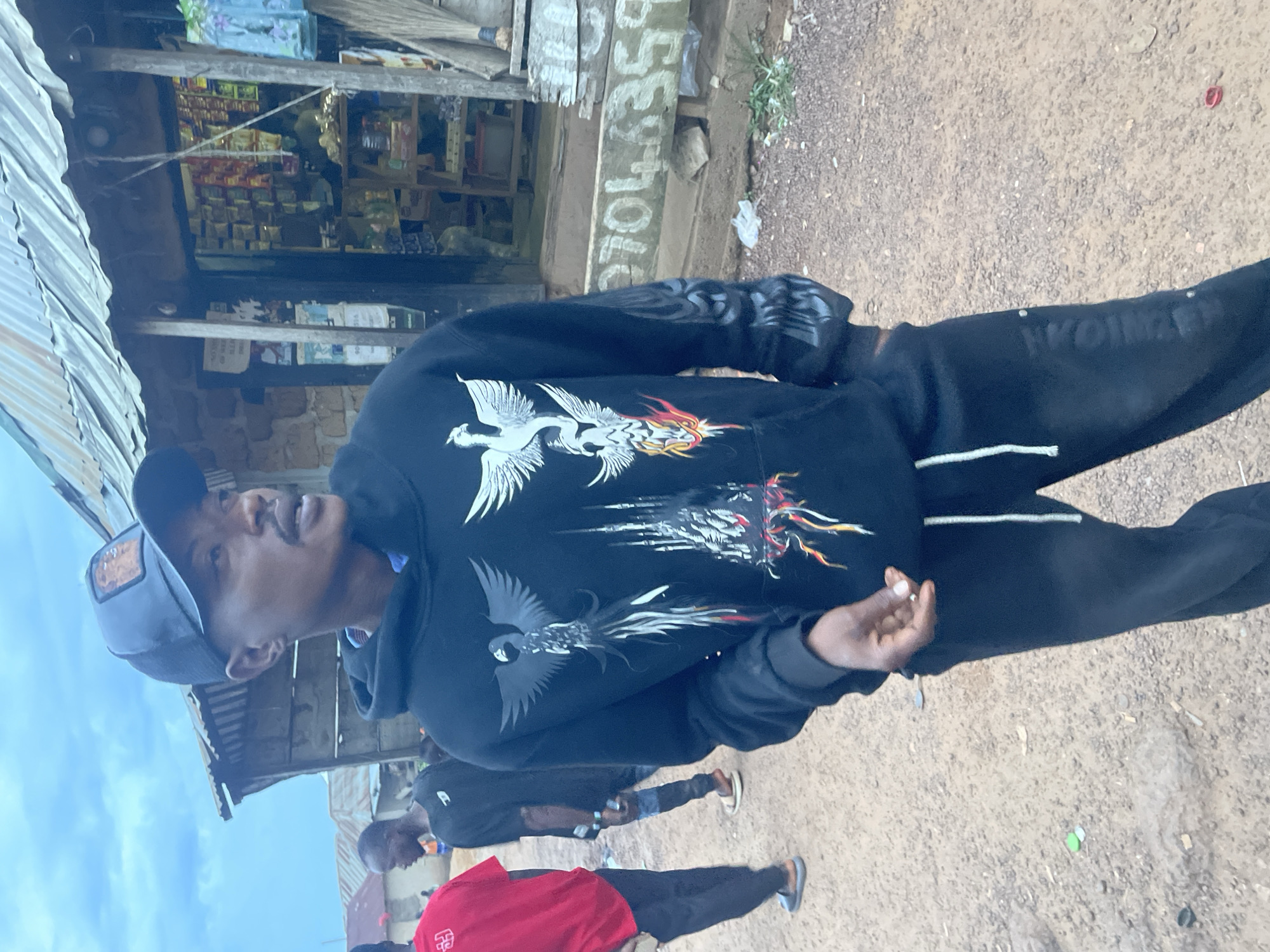A man wearing a black hoodie with a phoenix design, black sweatpants, and a blue baseball cap stands in an unpaved area of Durimi, Nigeria. He is facing left and slightly upwards. Two other people are walking behind him, one wearing a red shirt. A storefront with goods visible inside is behind the man. The ground is dirt and gravel. Some graffiti is visible on the side of the building.