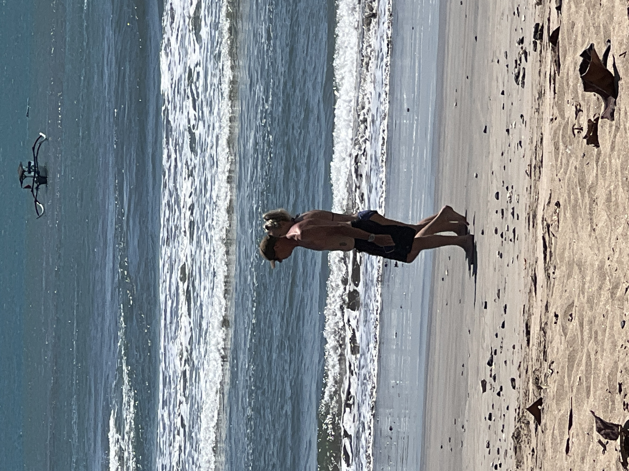 In Kuta, Indonesia, a couple walks along the shore of the ocean. The man, wearing a hat and dark shorts, is shirtless. The woman, in a bikini, walks beside him. The ocean has moderate waves, and a traditional Indonesian fishing boat is visible in the distance. The beach is sandy with scattered debris.