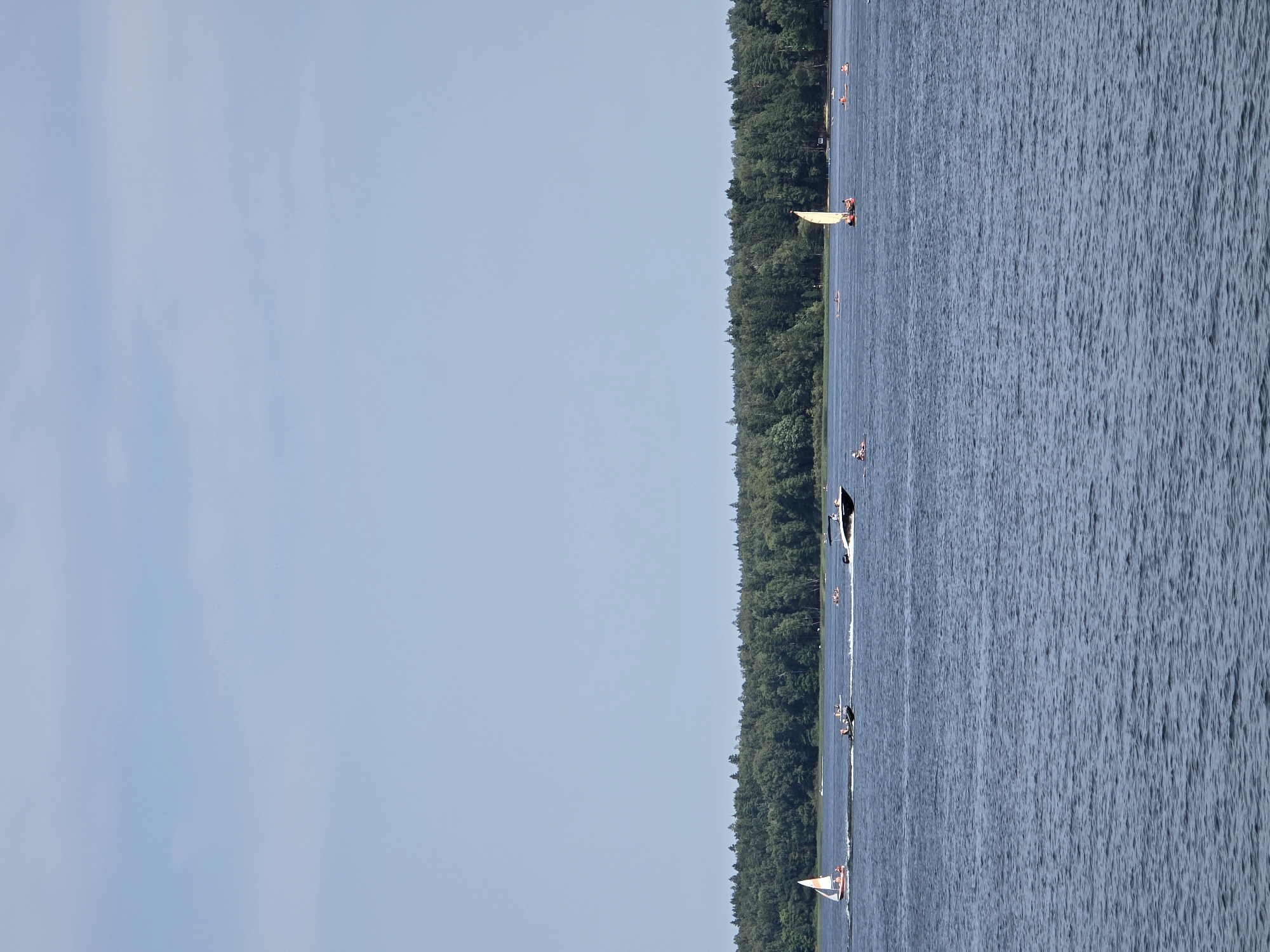 The image depicts a wide, calm body of water with a dense, green forest lining the far shore. The sky above is a clear, pale blue with some faint clouds. Several boats are visible on the water. On the left, a small sailboat with an orange-red sail is moving away from the viewer. Further to the right, a black speedboat with people on board is creating a white wake. In the middle distance, there are several other small boats or rafts with people on them, scattered across the water. Closer to the right side of the image, a larger sailboat with a light yellow sail is present, with at least two individuals on board. The overall scene suggests a recreational activity on a lake or large river. The location is identified as the посёлок гидроузла имени Куйбышева, Russia.