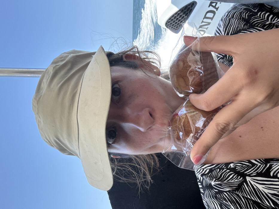 The image displays an adult individual situated on a motorized boat in Pak Nam, Thailand. The person, positioned from the chest up, wears a tan wide-brimmed hat and a black and white patterned top. Their dark eyes are wide open, directed towards the viewer. The individual holds two individually plastic-wrapped muffins in their left hand, with their mouth pressed against the clear plastic of the muffin on the left. A small, light-colored earring is visible in their right ear. The background features a clear blue sky meeting a body of blue water, with a white wake trailing behind, indicating the boat is actively moving. A silver metal pole of the boat structure rises on the right side of the frame, alongside a white boat panel featuring a black grille and the partially visible brand name "HONDA."