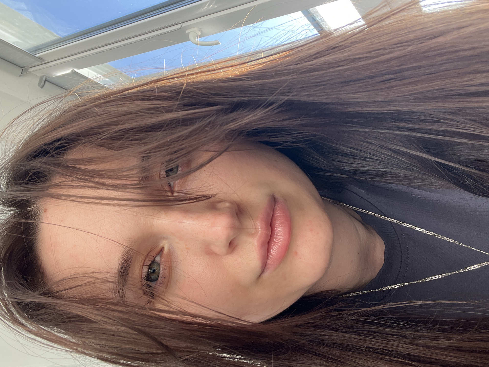 The image is a close-up, rotated portrait of a young person, likely female, with long, dark brown hair. The subject's head is tilted, and the image is oriented horizontally, giving the impression of the person lying down. They have light-colored eyes, possibly green or hazel, and a neutral to slightly pensive expression, with their lips gently parted. Strands of their hair frame their face, some falling across their left eye.
In the upper portion of the frame, a white ceiling and a skylight or ceiling window are visible. Through the window, a clear blue sky is seen, indicating bright, sunny daytime weather. White window frames and a mechanism, possibly for blinds or shutters, are present within the window structure. The subject is wearing a dark-colored t-shirt, and a thin silver chain necklace is visible against the fabric. The lighting is natural, suggesting it originates from the skylight. The setting is an indoor space in Tyumen, Russia. No text is visible in the media file.