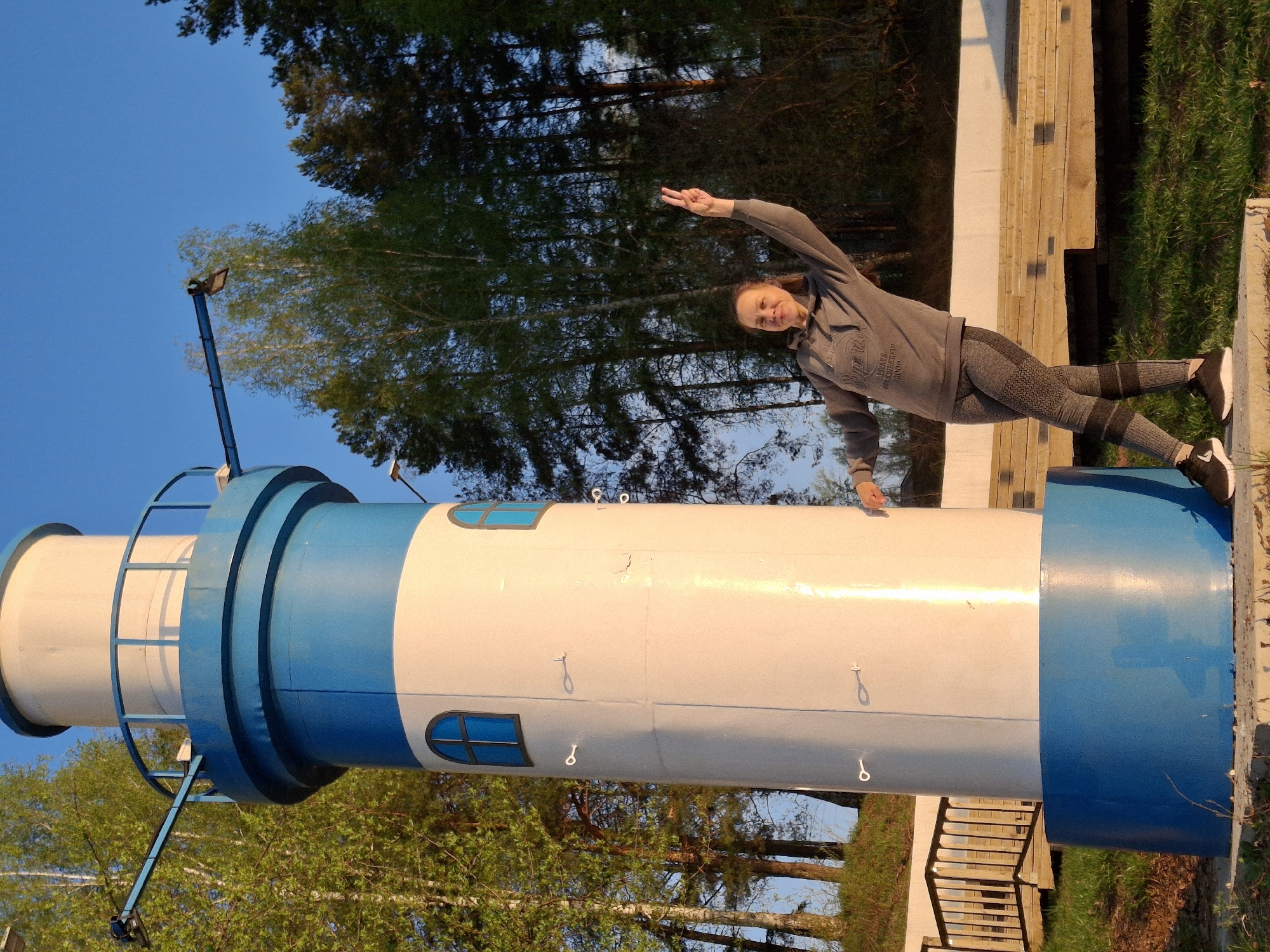 A young woman is posing in front of the lighthouse in Chaikovsky, Russia. The lighthouse is painted with blue and white bands and features small windows. The woman is wearing a gray hoodie and patterned leggings, striking a pose with one arm raised and making a peace sign. Trees and greenery are visible in the background. A boardwalk-style structure is to the right.