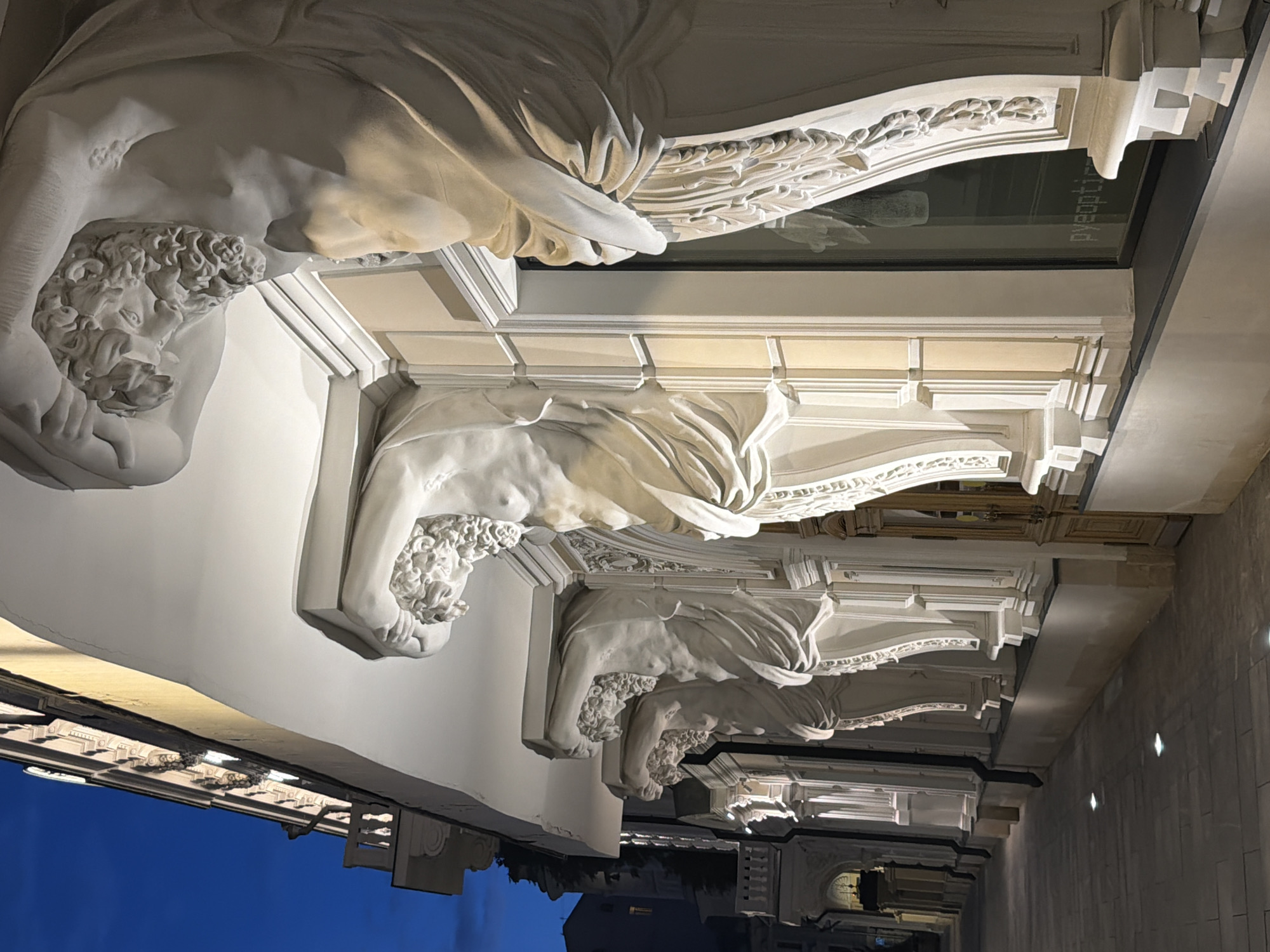 The image shows the illuminated facade of a building at twilight. A row of male atlantes sculptures, carved in white stone, are supporting the building's overhang. These figures are depicted with muscular torsos, draped clothing, and detailed facial features, including beards and curly hair. The atlantes are evenly spaced along the length of the building, each positioned to hold up a section of the protruding structure. Behind the atlantes, there are windows and architectural details of the building, illuminated by interior lights. To the left, a sidewalk paved with stone tiles is visible, with embedded lights casting a subtle glow on the ground. The sky above is a deep blue, indicating the late hour. The architectural style suggests a historical or classical influence. This building is located at 10 Tverskaya Street in Moscow, Russia, and the atlantes sculptures are a notable feature of its design.