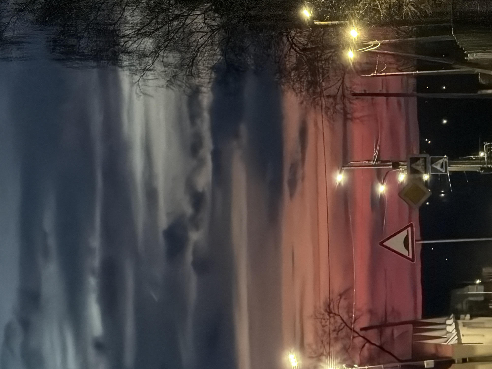 The image, when viewed correctly oriented, captures an outdoor scene in Chaikovsky, Russia, likely during twilight (either dusk or dawn). The sky dominates the upper portion, displaying a dramatic gradient of colors, transitioning from dark blues and grays on the left to vibrant pinks and oranges on the right, indicative of sunrise or sunset. Prominent, streaky clouds are visible across the sky.
Silhouetted bare tree branches frame the top left and central areas of the image. Numerous bright, warm-toned artificial lights, resembling streetlights or decorative illumination, are scattered throughout the scene, casting glows, particularly on the right side and towards the bottom.
In the mid-right section, two distinct road signs are visible: a yellow diamond-shaped sign and a white triangular warning sign with a red border, depicting a black wavy line, which typically indicates a bump or uneven road ahead. Indistinct dark structures or buildings can be seen on the far right.
The scene is static, devoid of people or specific activities, focusing on the atmospheric transition of light and the urban/suburban infrastructure under a dramatic sky. No visible text is discernable on the signs or elsewhere.
