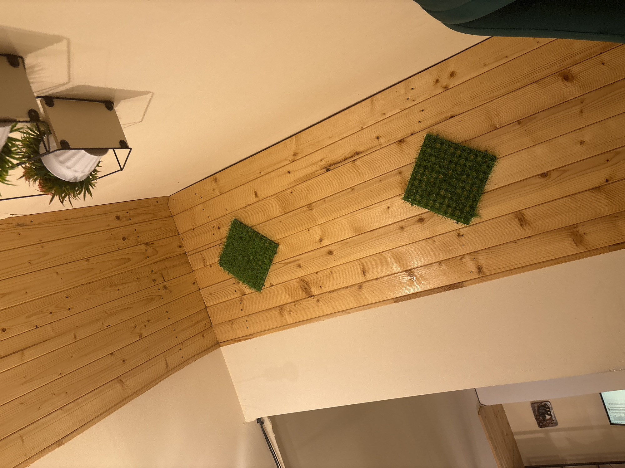This media file captures an indoor scene, likely a room or part of a building, characterized by its distinctive wall and ceiling treatments. The most prominent feature is a sloped surface, either a wall or ceiling, entirely clad in light-colored wooden planks arranged horizontally. These planks display natural wood grain and knots. Two small, square patches of artificial green grass are affixed to this wooden paneling; one is positioned towards the upper right, and the other slightly lower and to the left.
In the upper left portion of the image, a modern, multi-tiered planter or shelf is visible. It features a black metal framework holding white pots containing lush artificial plants with green and reddish-brown foliage. These plants cast shadows on the adjacent light-colored wall or ceiling. A glimpse of dark green upholstered material, possibly from a sofa or bed, is seen in the upper right corner.
The lower sections of the room consist of smooth, light-colored walls, appearing to be painted white or off-white. In the bottom right corner, several elements are partially visible: a dark, decorative ceiling light fixture with an ornate design, a vertical wooden beam or post, and a white rectangular object that resembles a screen or monitor with faint graphical display.
The scene is static, with no discernible activity or people present. The lighting suggests artificial indoor illumination. No text is visible within the image. The overall impression is a contemporary interior with natural and artificial elements, located in Kardzhali, Bulgaria.