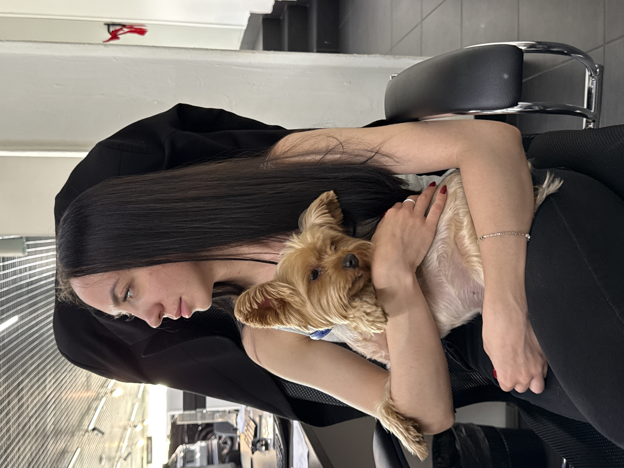 The image shows a young woman holding a small, light brown dog. She has long, dark brown hair and is wearing a black top and black leggings. A thin, sparkly bracelet is visible on her left wrist. The dog appears to be a Yorkshire Terrier or a similar breed. The woman is cradling the dog in her arms, and they are both looking downwards and to the left. The scene appears to be indoors, possibly in a salon or spa, given the background elements of sleek chairs and what looks like hair salon equipment. The lighting is bright, suggesting it's daytime. The overall emotion conveyed is one of tenderness and affection between the woman and her dog. No visible text is present in the image.