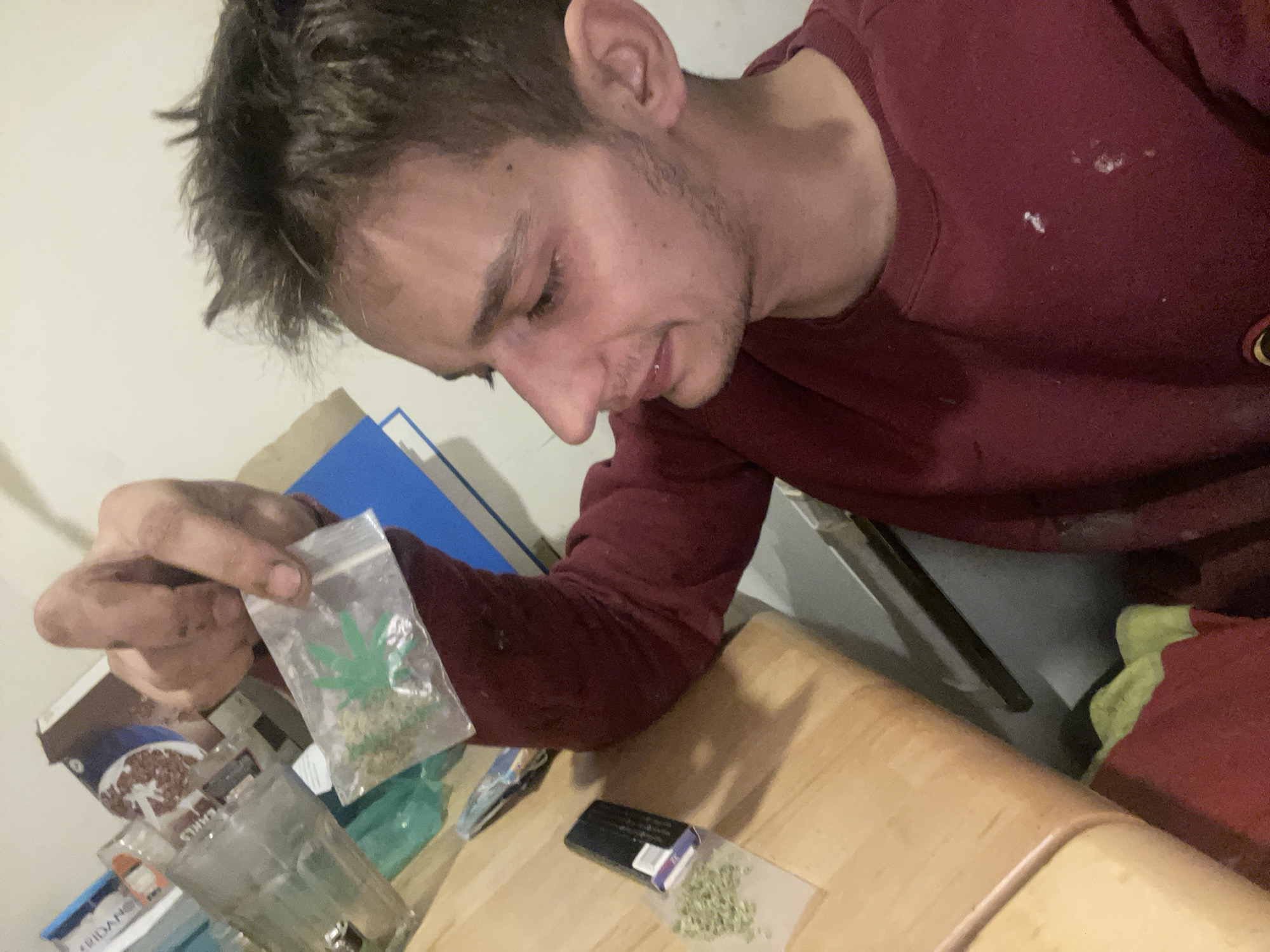In an indoor environment with a light-colored wall and a wooden table, a young Caucasian male with dark, messy hair and stubble is the central subject. He is wearing a maroon sweatshirt, which has some white specks on it. The male is leaning forward, looking down at his hands. In his left hand, he is holding a small, clear plastic bag containing a green, leafy substance, possibly marijuana, with a green cannabis leaf design printed on it. To his right, on the wooden table, is another small, clear plastic bag with a similar green, leafy substance, and a black rectangular object with some white and purple markings. Behind the male, on the table, are several items including what appears to be a glass, a cardboard box with branding that includes images of cereal, and a blue folder. The overall scene suggests a casual, possibly private setting.