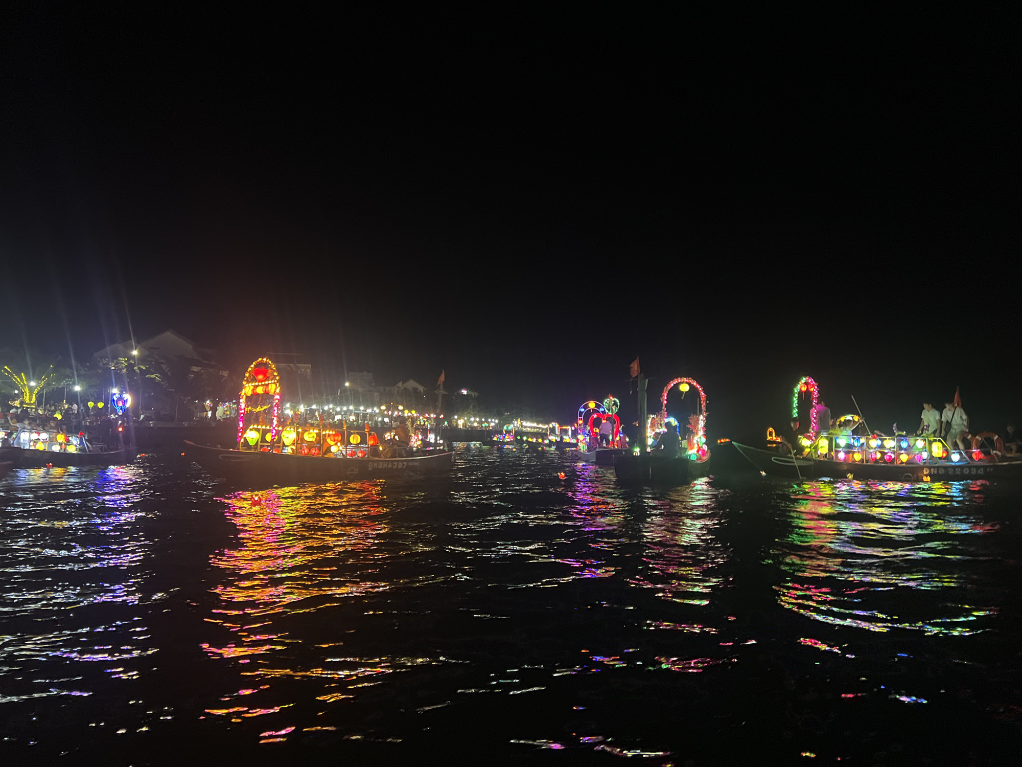 The image depicts a nighttime scene of a river in Hoi An, Vietnam, likely during a festival or celebration. Several boats are illuminated with colorful lanterns and decorations, casting vibrant reflections on the dark water. The boats are adorned with various lantern arrangements, including arches of lights and strings of hanging lanterns in shades of red, yellow, green, and blue. People are visible on some of the boats, engaged in activities related to the celebration. The riverbank in the background is also lit up with lights from buildings and trees, creating a festive atmosphere. The overall scene conveys a sense of enchantment and merriment.