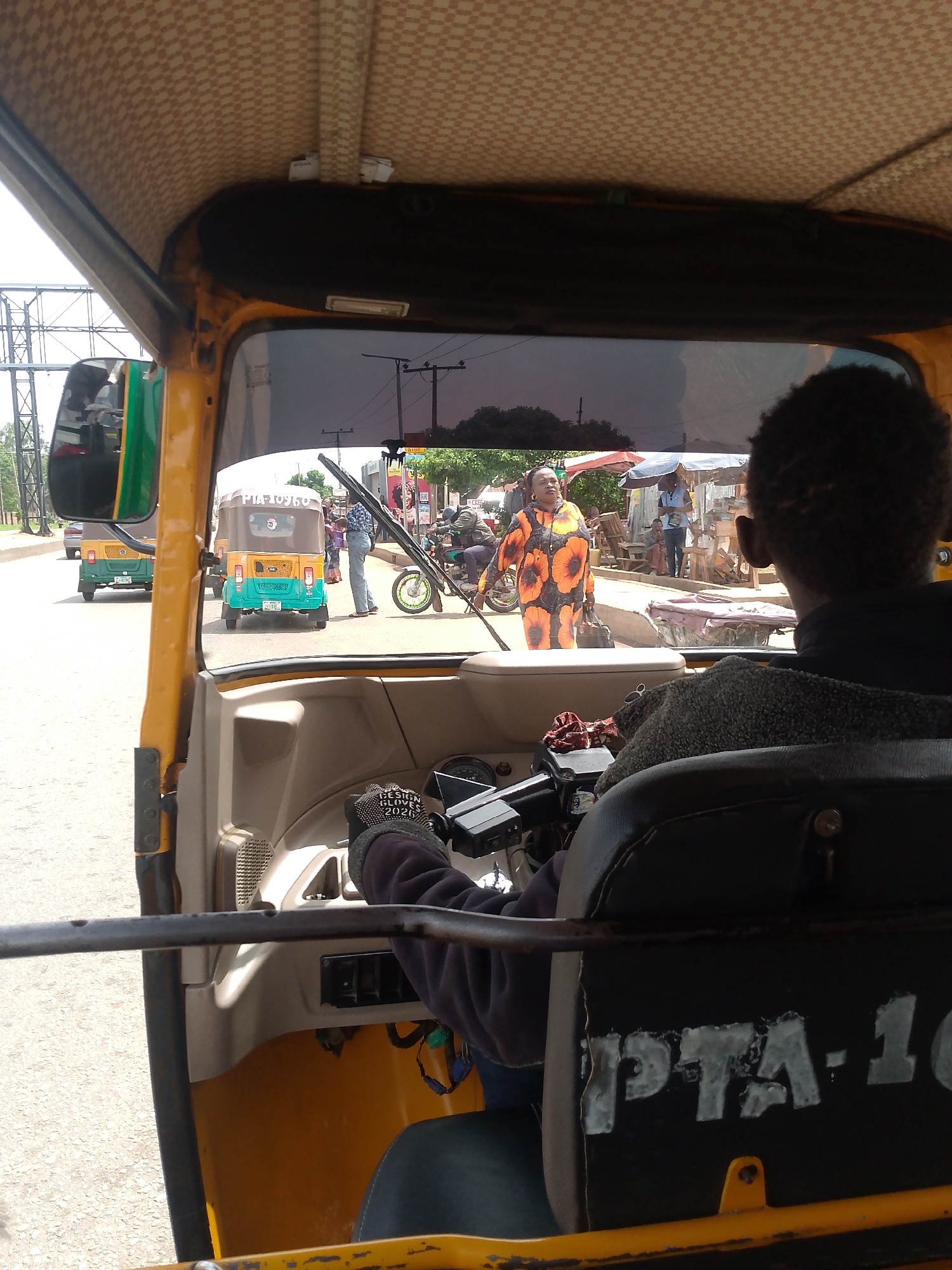 A view from inside a yellow and beige tuk-tuk in Bukuru, Nigeria. The driver, with dark hair, is visible from the back, wearing a dark sweater and gloves with writing on them. Through the front window, a woman in an orange dress with large sunflower-like designs is crossing the street. Other tuk-tuks, pedestrians, and a motorcycle are visible on the roadside with stalls and umbrellas. The sign PTA-10 is visible on the back of the driver's seat.