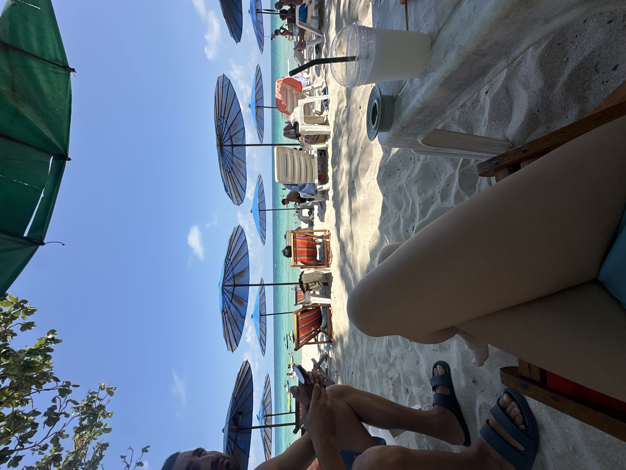 This image captures a beach scene in Baan Chaweng, Thailand, taken from a low angle. The sky is blue, with beach umbrellas lining the shore. Various beachgoers are visible, some lounging in beach chairs. In the foreground, a person sits in a chair wearing blue sandals. A drink with a straw sits on a table next to them.