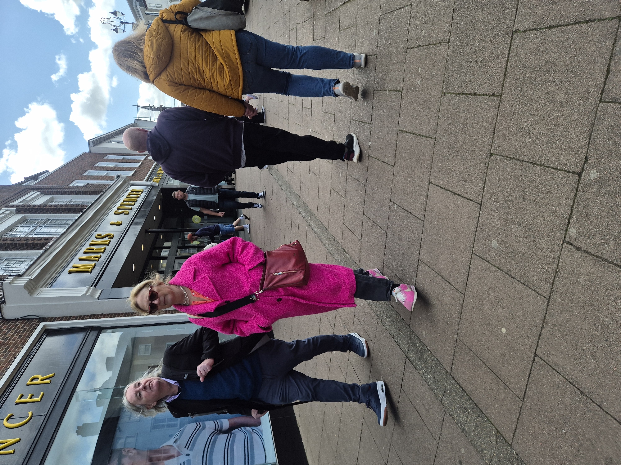 The image depicts a street scene in West Suffolk, United Kingdom. Four people are walking on a sidewalk. The storefront of "Marks & Spencer" is visible. A woman in a bright pink coat is in the foreground, carrying a red bag. She is wearing sunglasses and pink sneakers. A man in a dark jacket and blue shirt is walking next to her. An older man is reflected in the store window. A person wearing a yellow jacket and jeans is further away, as is a person with a bald head in a dark jacket and black trousers. The sky is blue with some clouds.