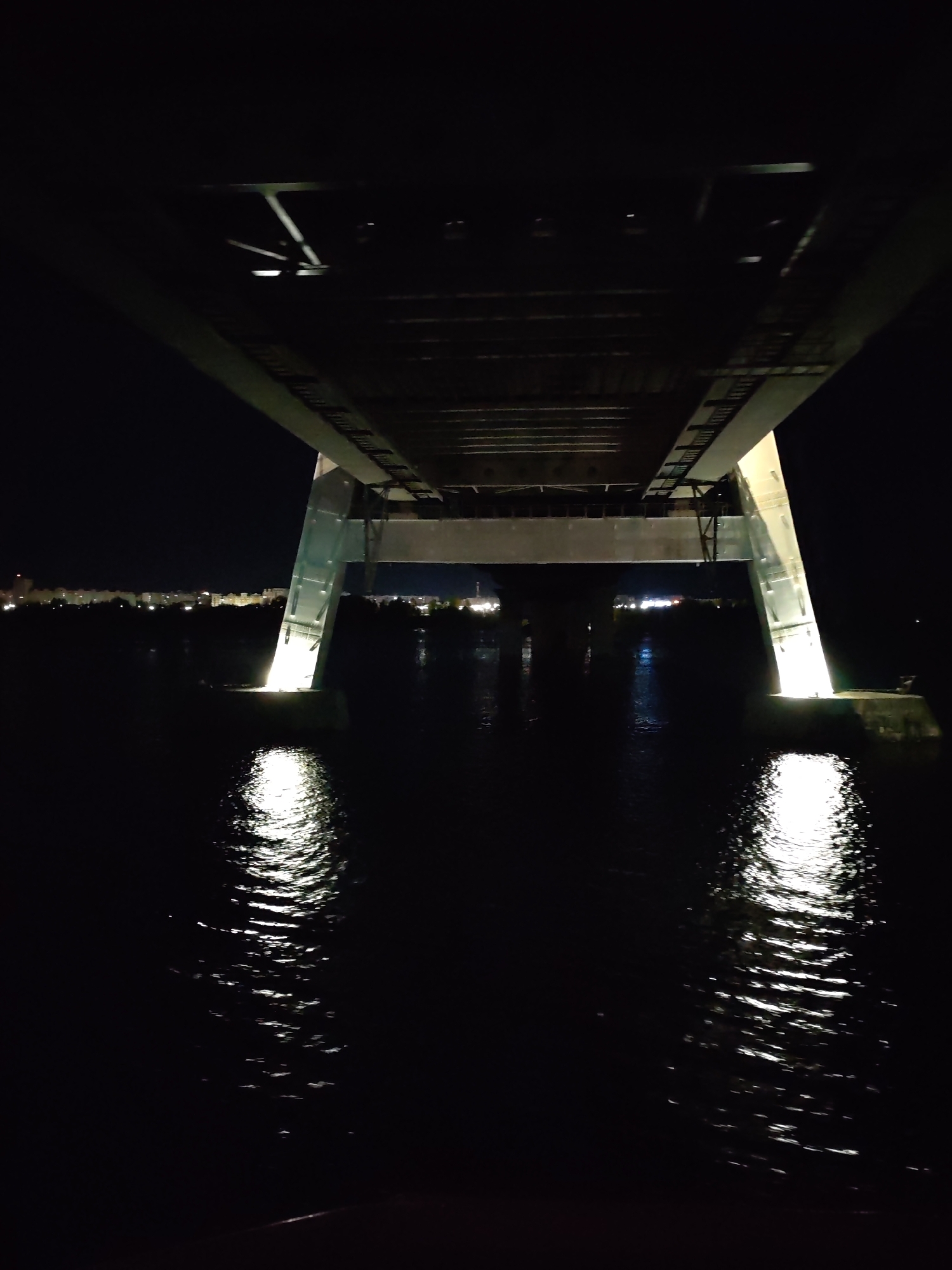A low-angle, night shot shows the underside of a bridge spanning a body of water. The bridge support columns, angled and illuminated, cast bright reflections on the water’s surface. In the background, faint city lights are visible. The location is in Cherepovets, Russia.