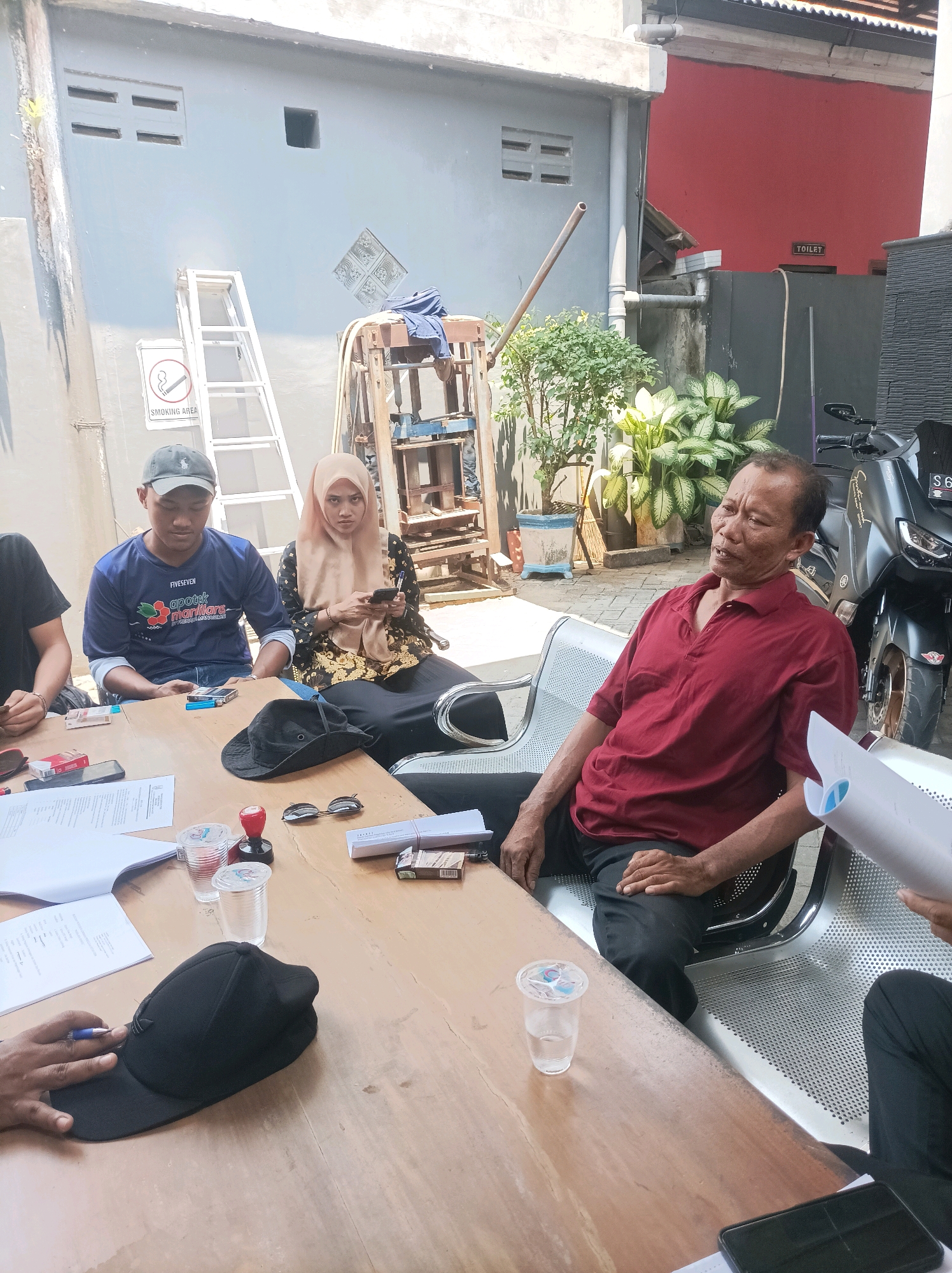 In the city of Rengel, Indonesia, a group of individuals are gathered around a wooden table outdoors. The scene appears to be in a semi-industrial or commercial area, with a gray wall and a red wall visible in the background, along with a motorcycle.
There are at least four people visible. A man wearing a dark blue t-shirt with white text and a dark baseball cap is seated at the table, looking down. Beside him, a woman wearing a light brown hijab and a black and gold patterned dress is seated, looking at her phone. To their right, an older man in a red collared shirt is seated, leaning back and looking to the side. Another person's arm and hand, holding a pen and wearing a black glove, are visible on the left side of the frame, reaching towards a black baseball cap on the table.
On the table, there are several items: papers, pens, glasses with liquid, a red stamp with a black base, sunglasses, a black baseball cap, and packs of cigarettes. A ladder and a "Smoking Area" sign are visible on the gray wall behind the people. The overall atmosphere suggests a meeting or discussion taking place.
