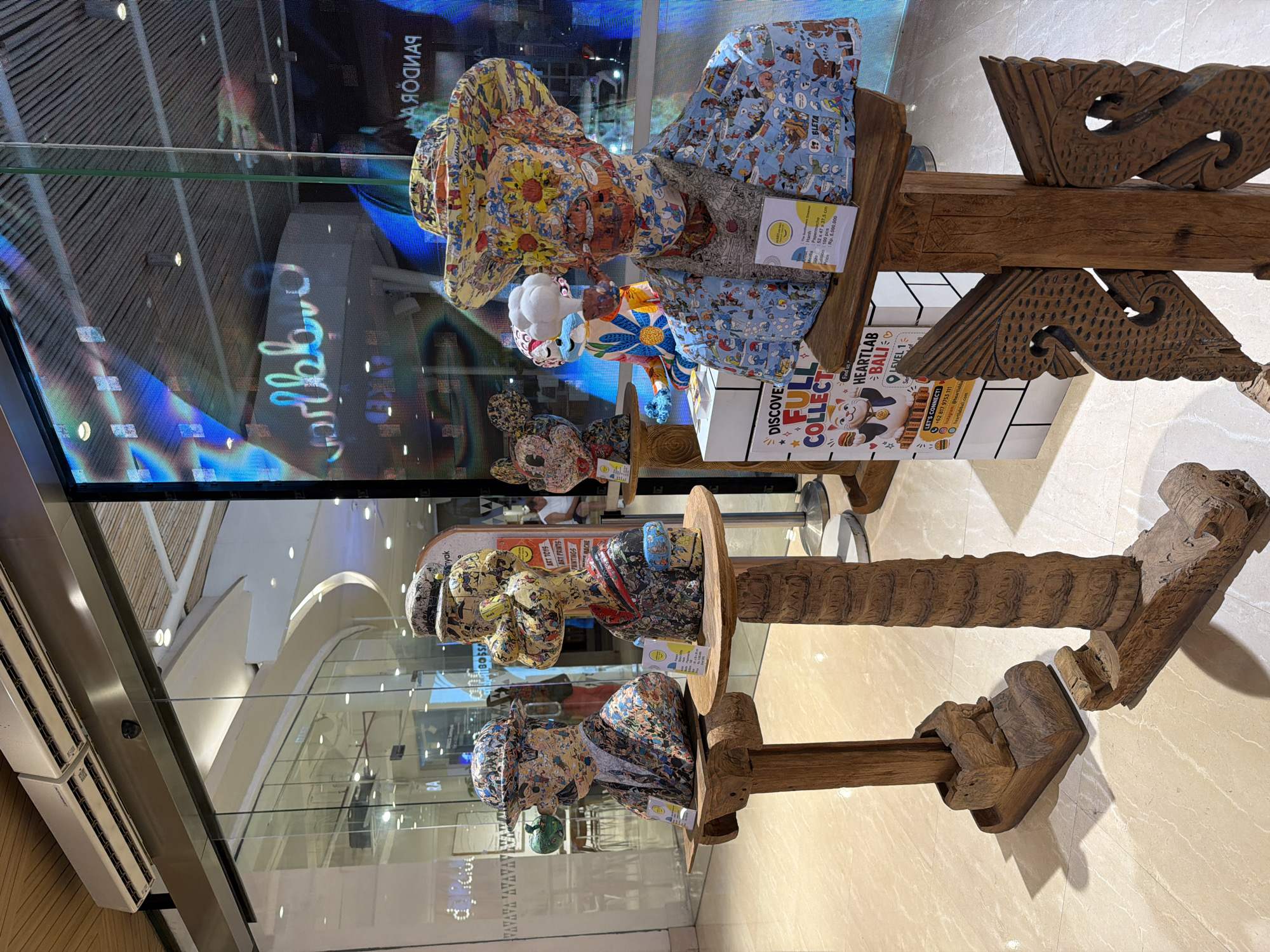 Here is a description of the image:
The photo shows three wooden pedestals, each supporting a papier-mâché-like figure. The figures appear to be artistic depictions of cartoon characters. The base of the pedestal has a card with the words "HEARTLAB BALI" and "LEVEL 1" which is a store in Kuta, Indonesia. The figures are behind a glass window. Reflections in the glass indicate the presence of an advertisement featuring "PANDORA." The floor beneath the figures is a light-colored tile.
