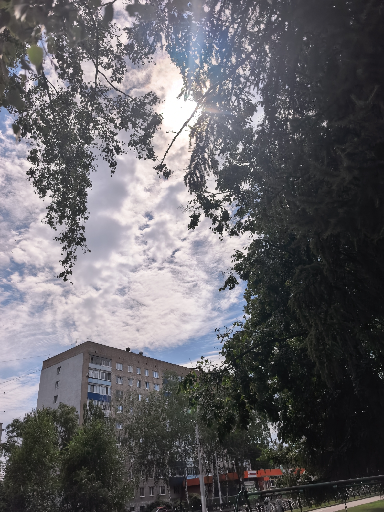 A low-angle shot captures a cloudy sky above a residential building in Sterlitamak, Russia. The building, with its brick and white facade, is partially obscured by the branches of trees in the foreground. Sunlight filters through the leaves and clouds, creating a dappled effect. The scene appears to be a typical urban environment with visible power lines and streetlights. No people or specific activities are depicted.