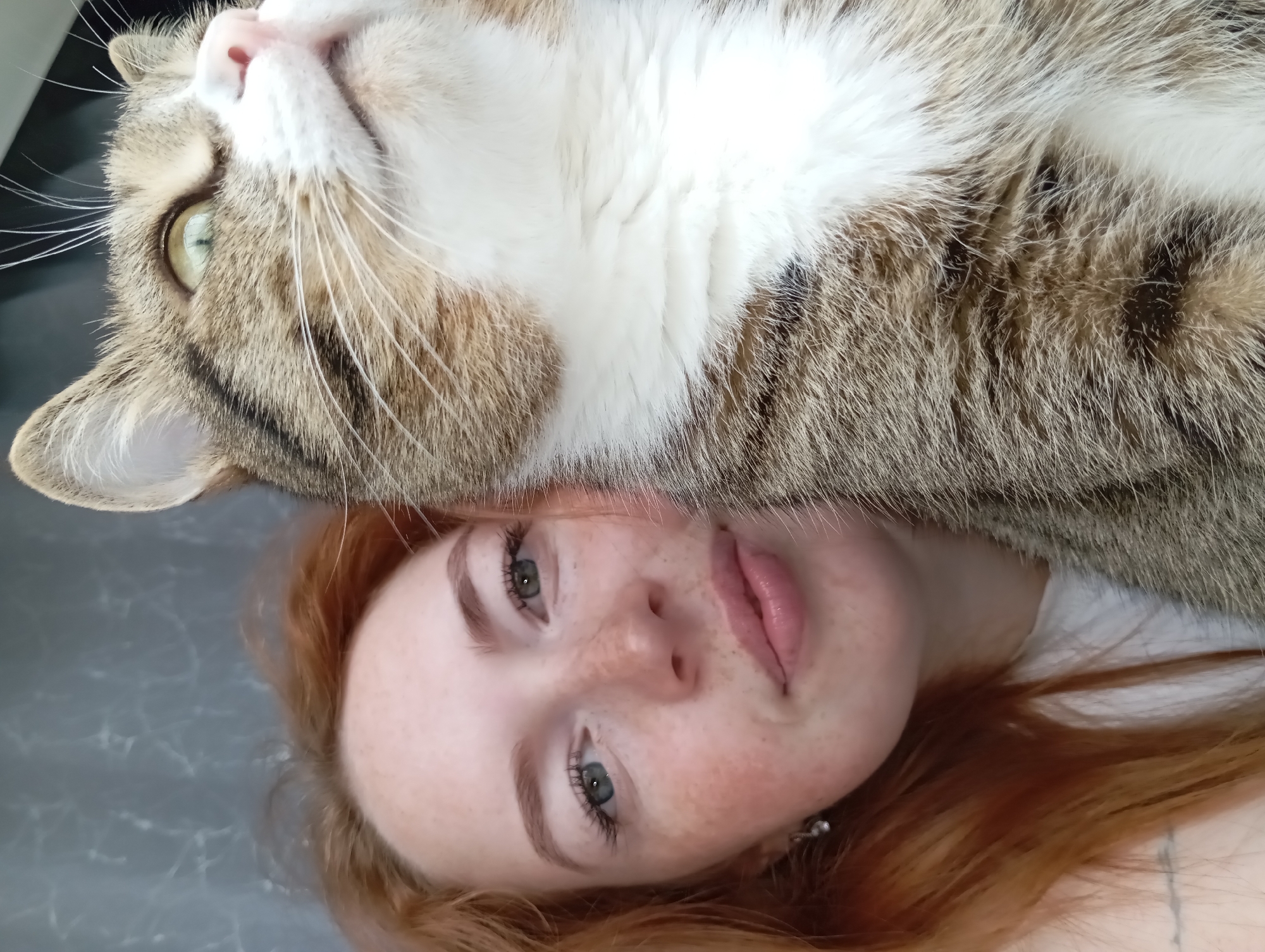 A human female with red hair, freckles, and blue eyes is lying on her back, with a tabby cat lying on top of her. The cat is facing towards the left, and the woman is facing towards the right. The location appears to be indoors. There is no indication that the location is in the city of Yurgamysh, Russia.