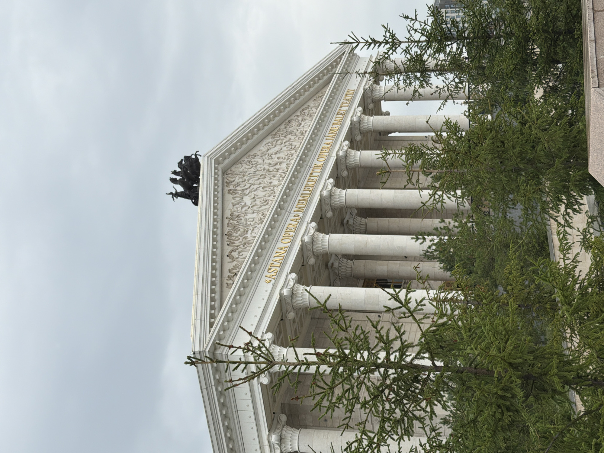 The image captures the Astana Opera House in Astana, Kazakhstan, under an overcast sky. The building is a grand neoclassical structure with a prominent white facade, adorned with Corinthian columns supporting a pediment. The pediment itself is decorated with intricate carvings, and atop it sits a bronze sculpture of a quadriga, a chariot drawn by four horses, with a figure standing in it.
In the foreground, several green evergreen trees partially obscure the view of the building, their branches reaching upwards. The trees are dense with needles and appear to be coniferous, possibly fir or spruce trees. The ground in front of the trees is not clearly visible, but a stone structure, possibly a low wall or planter, is present at the bottom of the frame. In the background, beyond the Opera House, a modern high-rise building with visible windows can be seen, indicating the urban setting of the location. The overall impression is of a majestic building set against a muted, cloudy day, with the trees adding a natural element to the scene. There are no people visible in the image. The text on the building reads "ASTANA OPERA «MEMLEKETTIK OPERA JANE BALET TEATRY»".