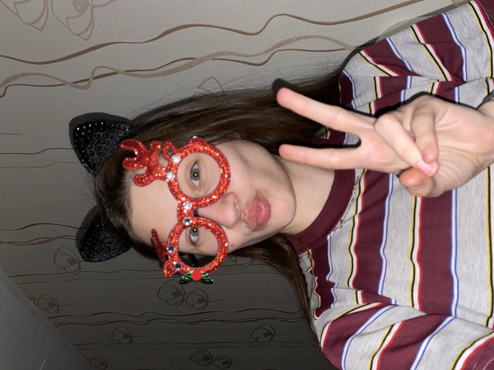 A close-up portrait shows a young person, with the image rotated 90 degrees clockwise. The person, with long brown hair, is looking directly at the camera while making a peace sign (V-sign) with their right hand. Their lips are puckered in a playful "duck face" expression.
They are wearing festive accessories: bright red glitter glasses shaped like reindeer antlers, adorned with small green holly details, and a dark, glittery headband with cat ears. The person is dressed in a striped t-shirt featuring vertical bands of grey, burgundy, white, blue, and yellow.
The background is an indoor wall covered with light-colored wallpaper. The wallpaper has a subtle pattern of wavy lines and abstract, possibly floral or stylized eye-like designs, some of which exhibit a sparkling or glittery texture. The overall impression is one of fun and celebration, possibly at a party. The location context provided for this image is Votkinsk, Russia. No visible text is present.
